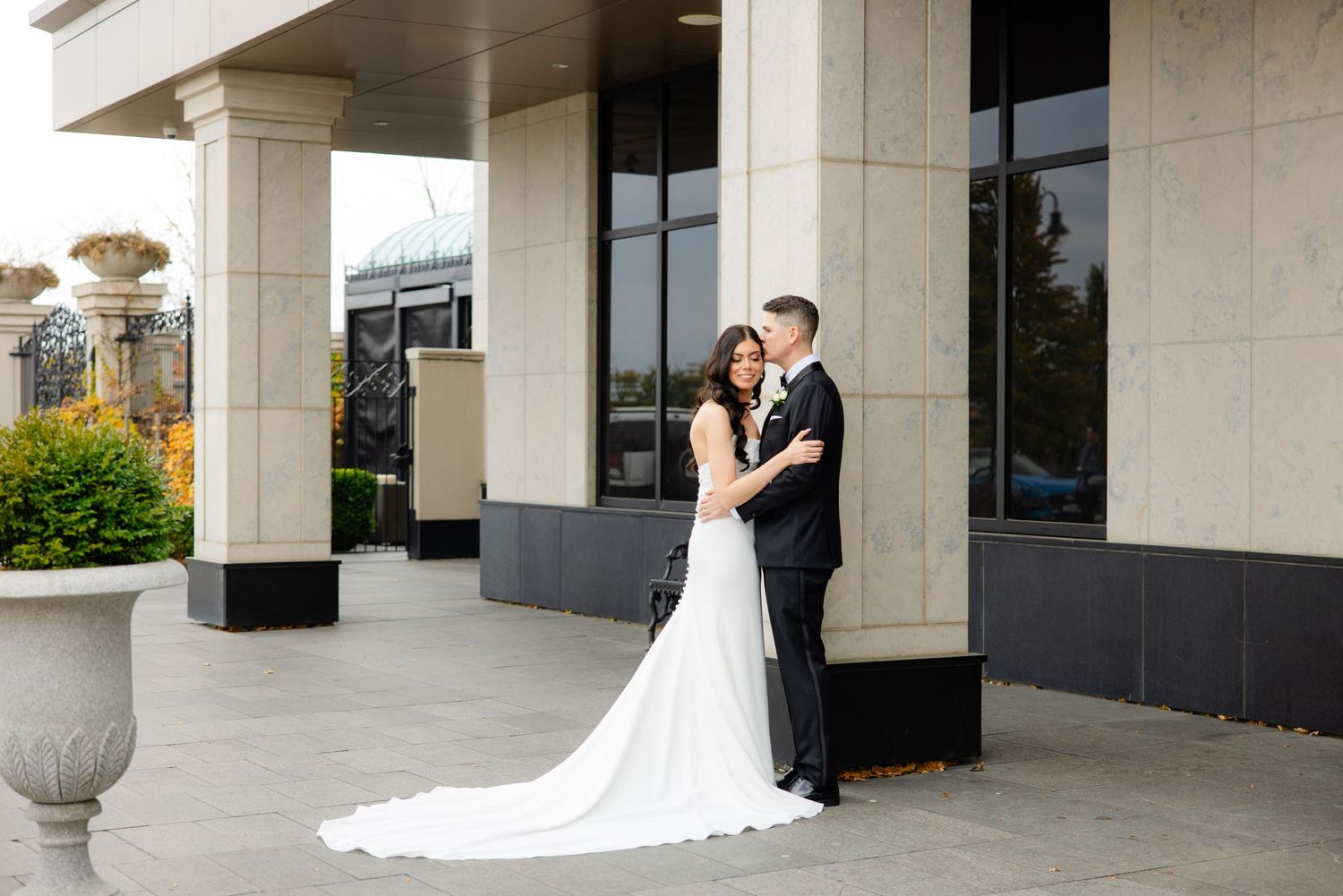 Intimate moment between newlyweds captured under building columns with long white train visible.