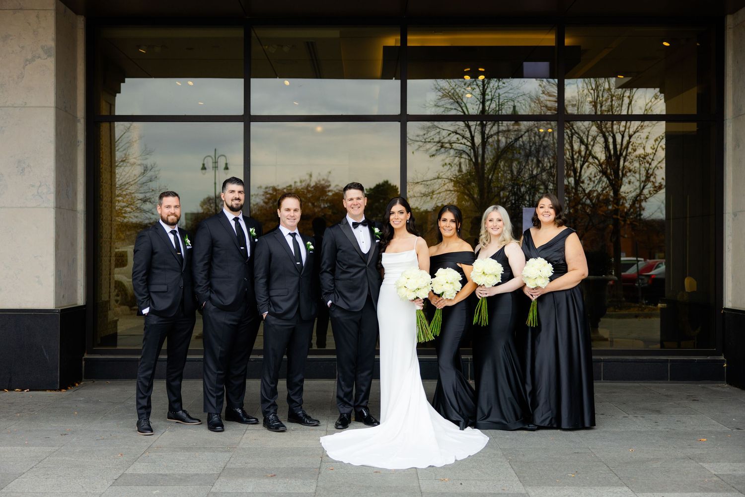 A formal wedding party poses together in navy and black attire outside a modern building.