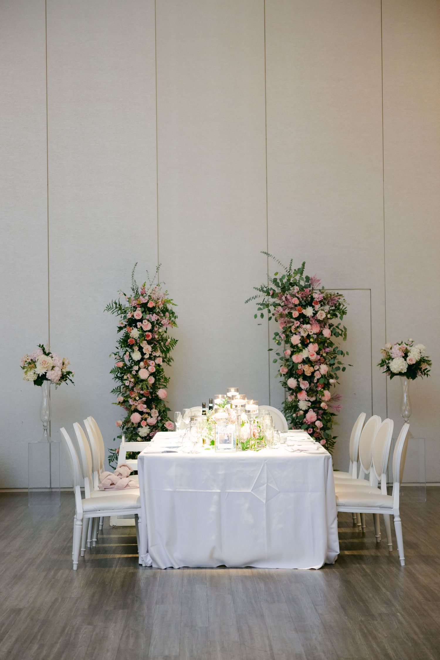Wedding head table setup with tall floral arrangements and white chairs against neutral wall.