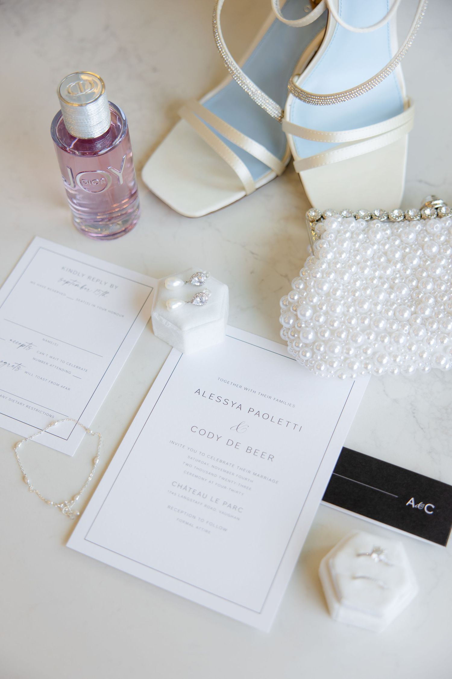 Wedding accessories including white heels, pearl clutch, and invitation with pink perfume bottle.