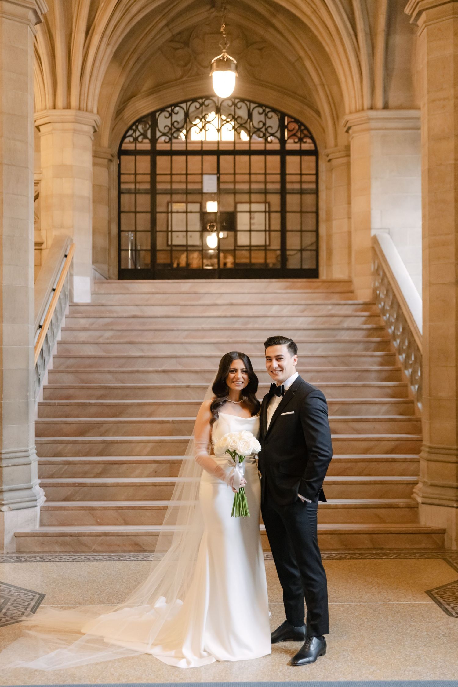A couple in wedding attire poses on grand marble steps with ornate architectural details.