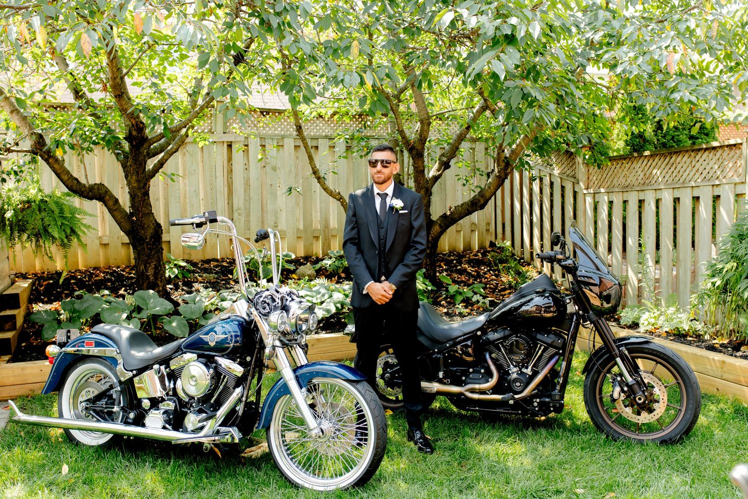 Suited figure standing with two motorcycles in a backyard garden setting with trees and wooden fence.