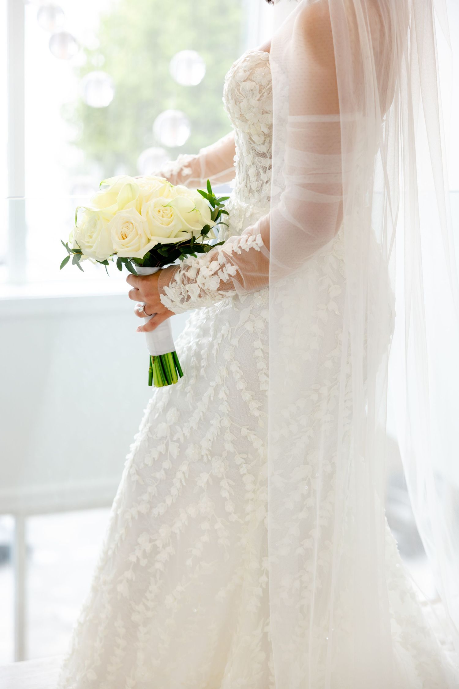Close-up of a bride wearing a detailed lace gown holding a bouquet of white roses near a window.