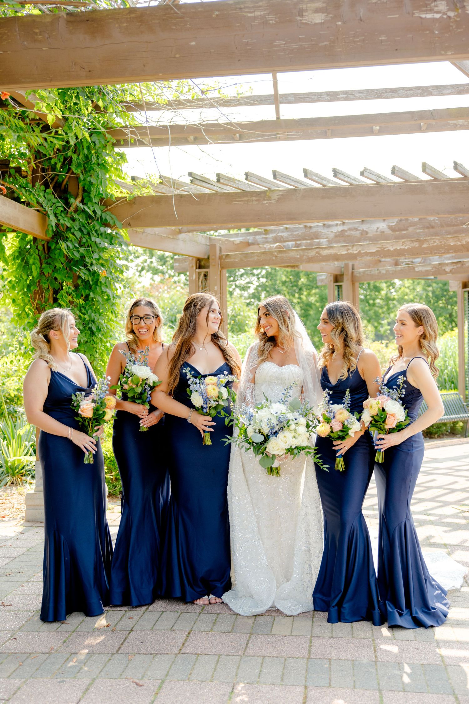 Wedding party laughs together under rustic pergola with bridesmaids wearing matching navy gowns.