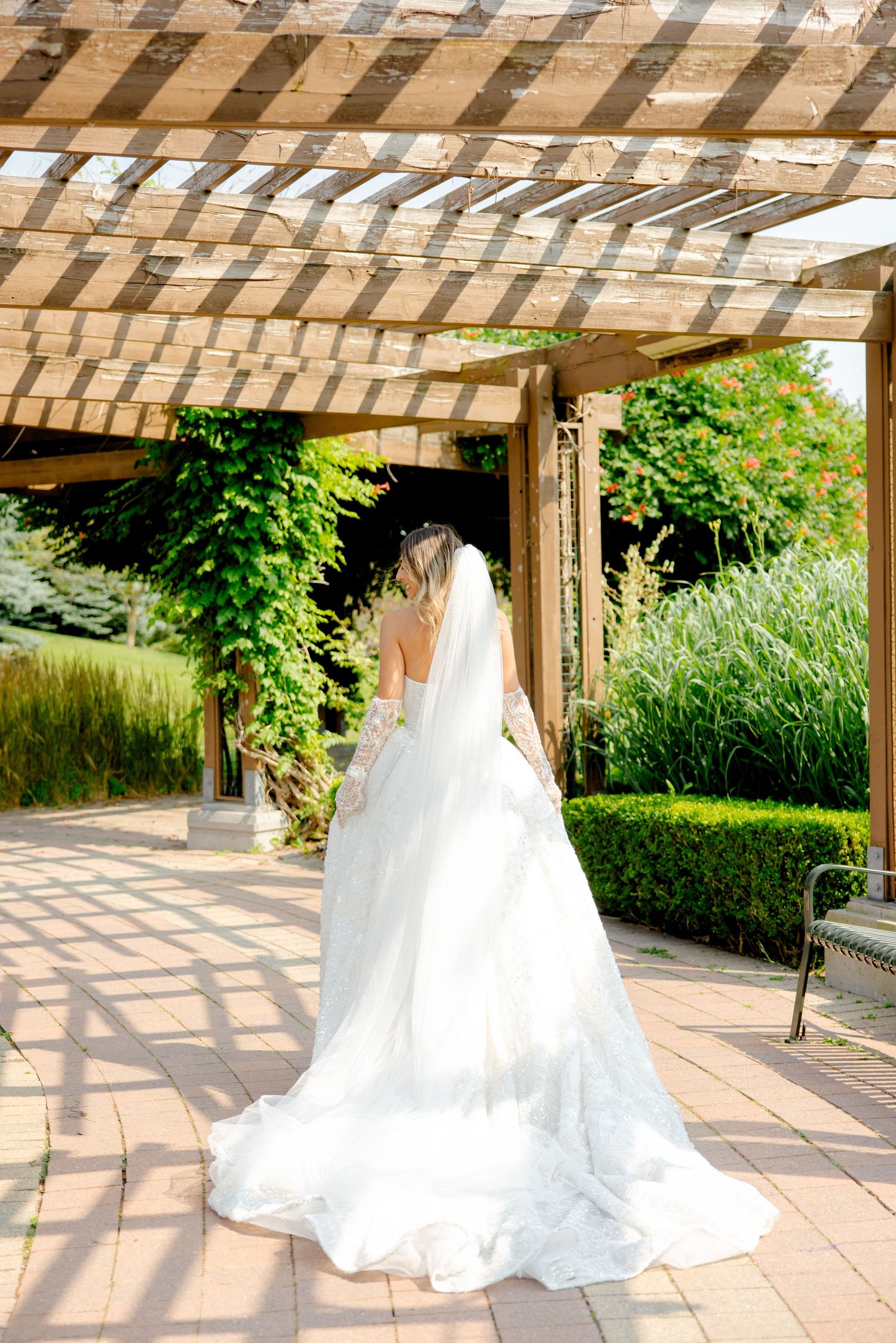 White wedding dress train flows on stone path under wooden pergola.