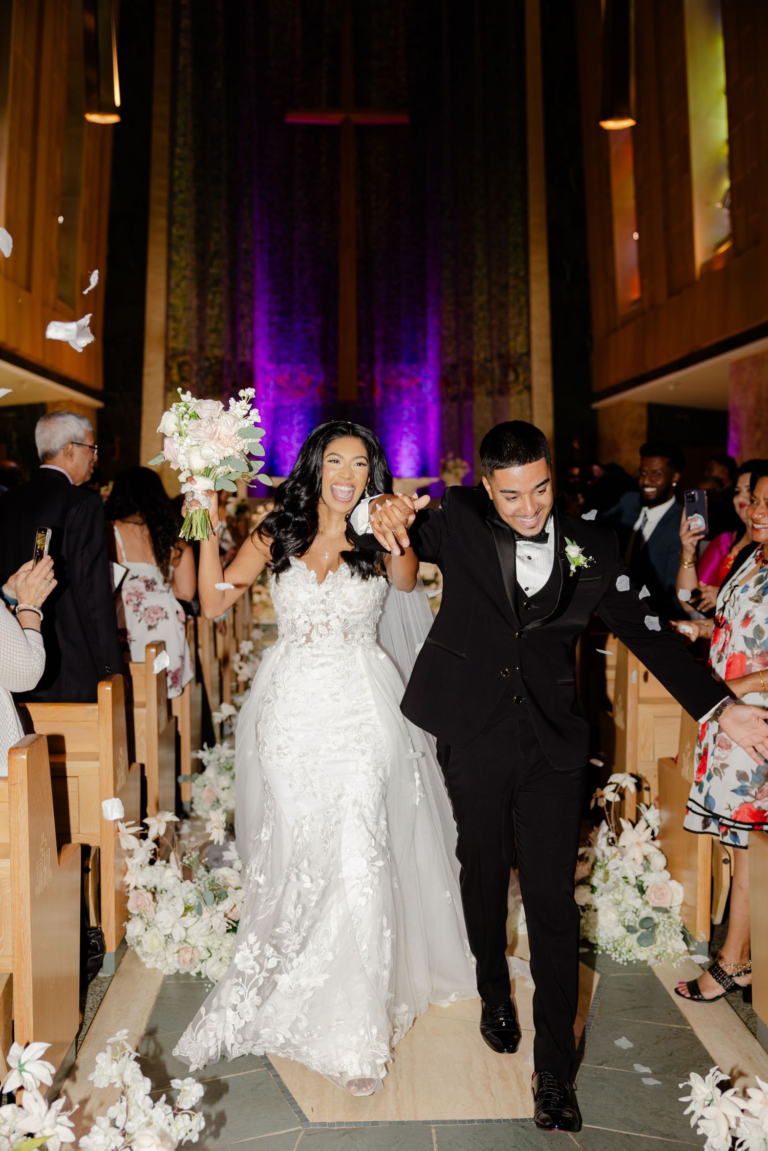 A wedding couple walks down an aisle lined with white floral arrangements in a church.