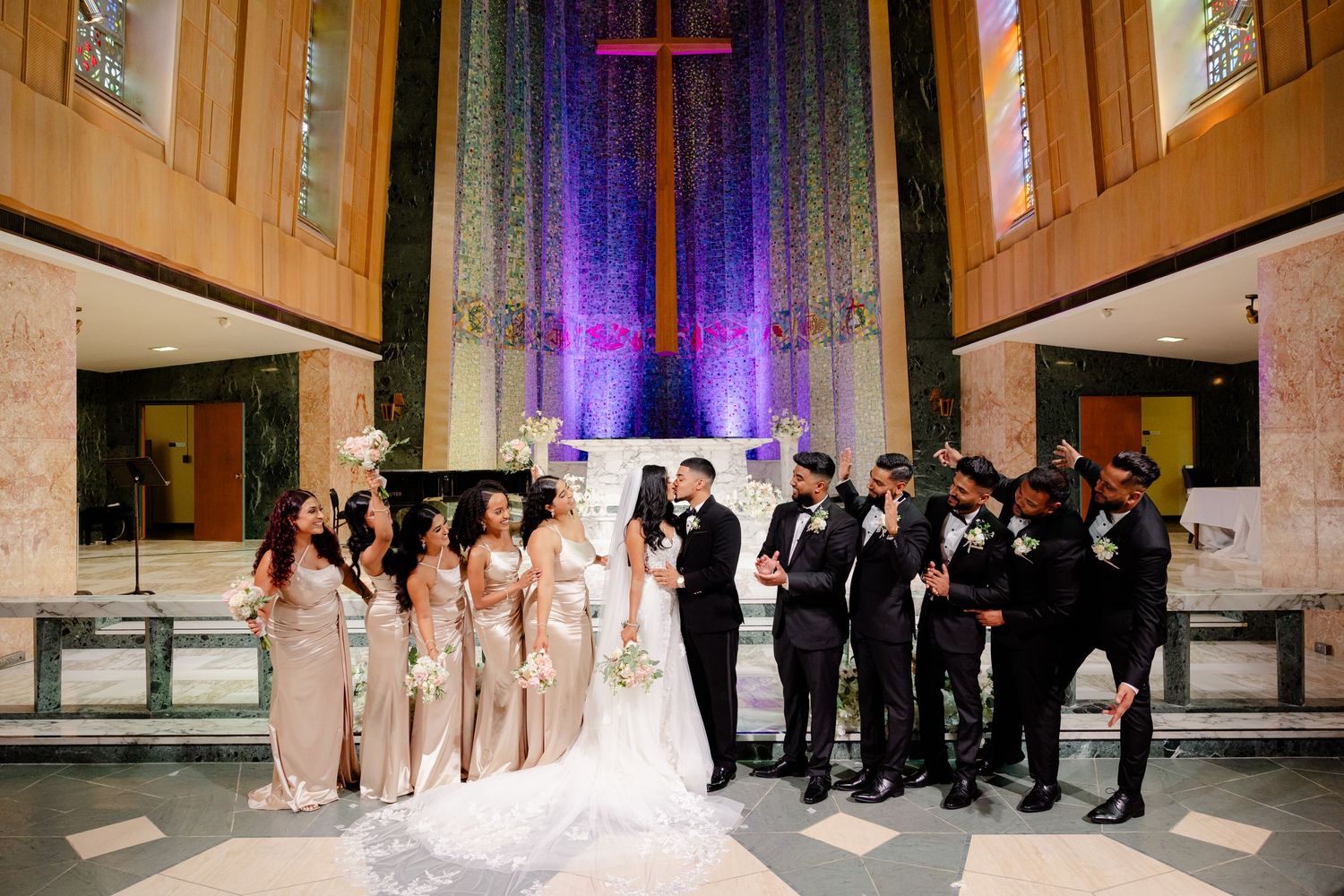 Formal wedding party photo with bridesmaids and groomsmen flanking the couple at church altar.