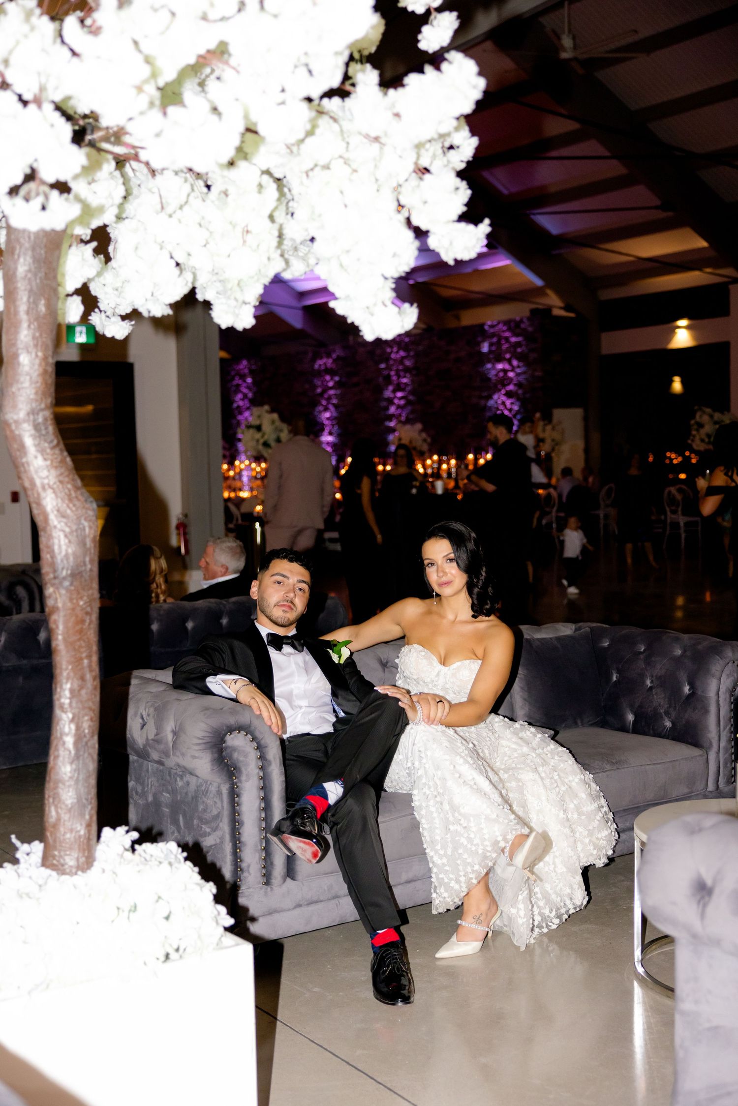 Newlyweds sharing relaxed moment on plush gray couch under decorative white tree.
