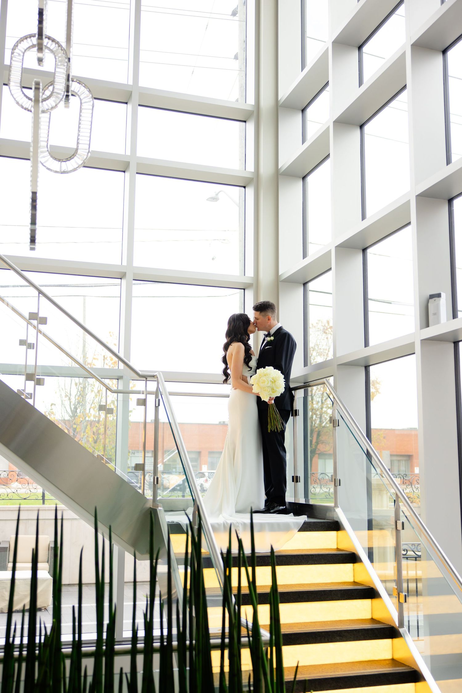 Elegant couple stands on modern yellow staircase surrounded by tall windows and architectural plants.