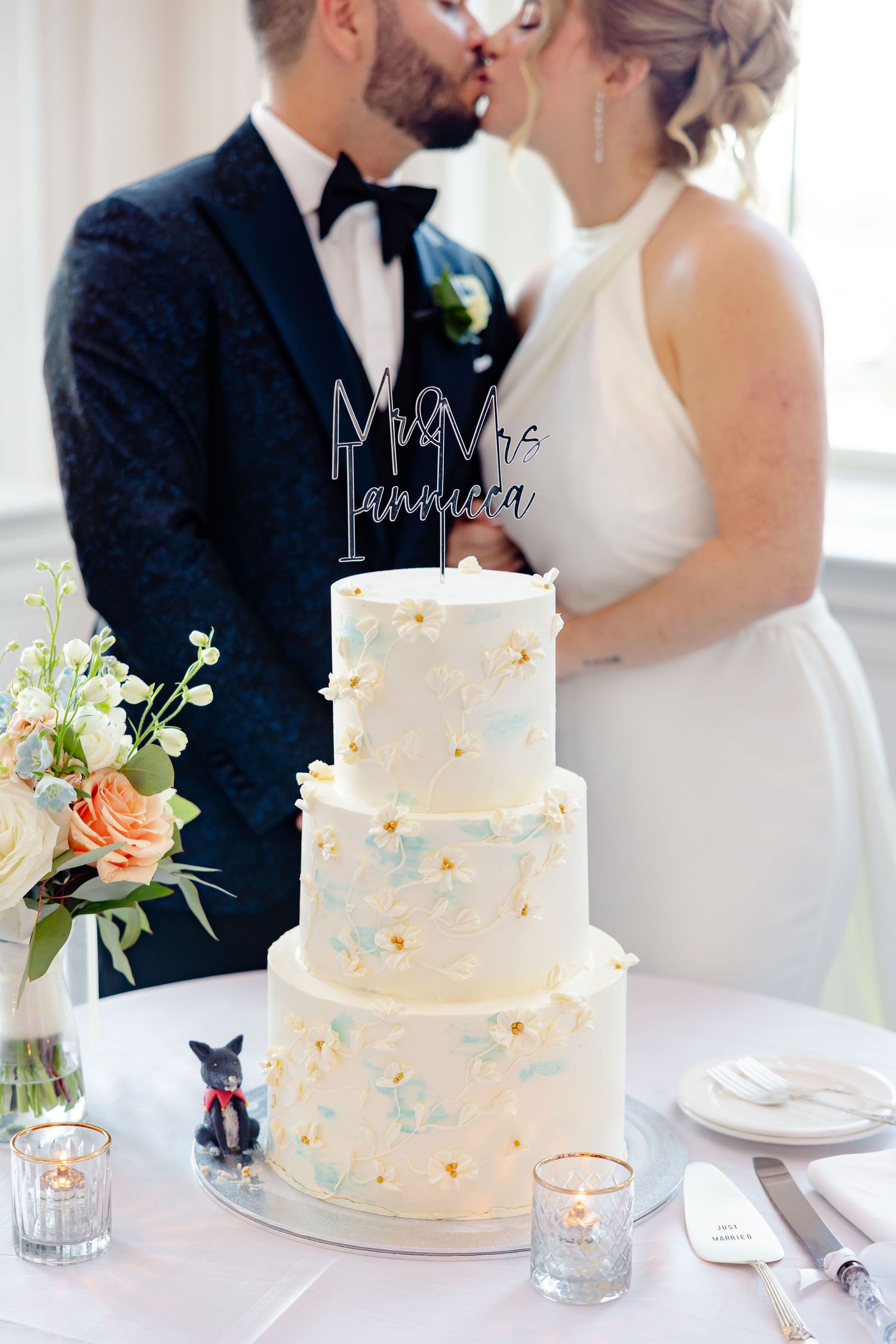 Newlyweds share a tender moment over their three-tiered white wedding cake adorned with delicate floral decorations.