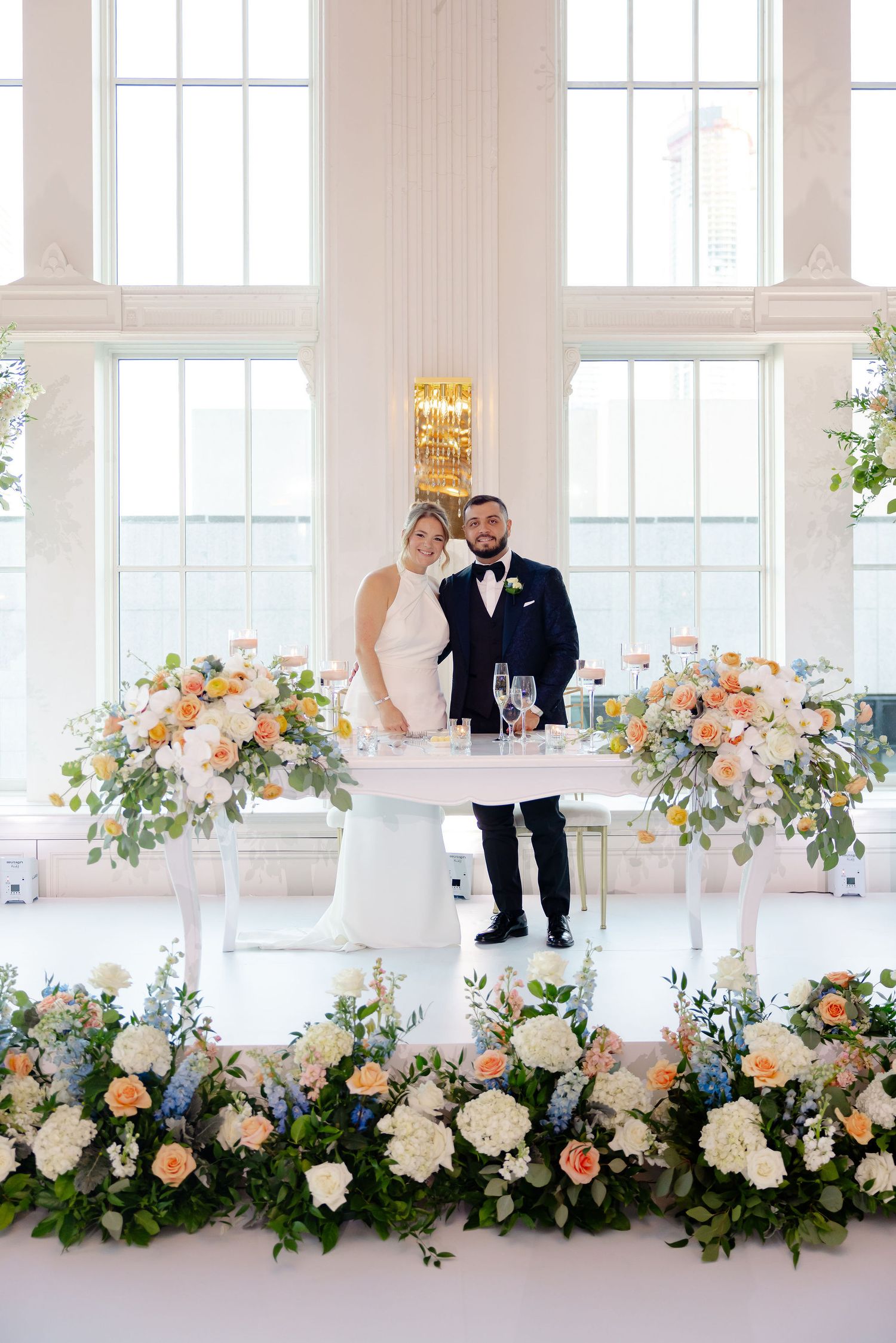 Head table decorated with lush peach and white floral arrangements in an elegant white reception room.