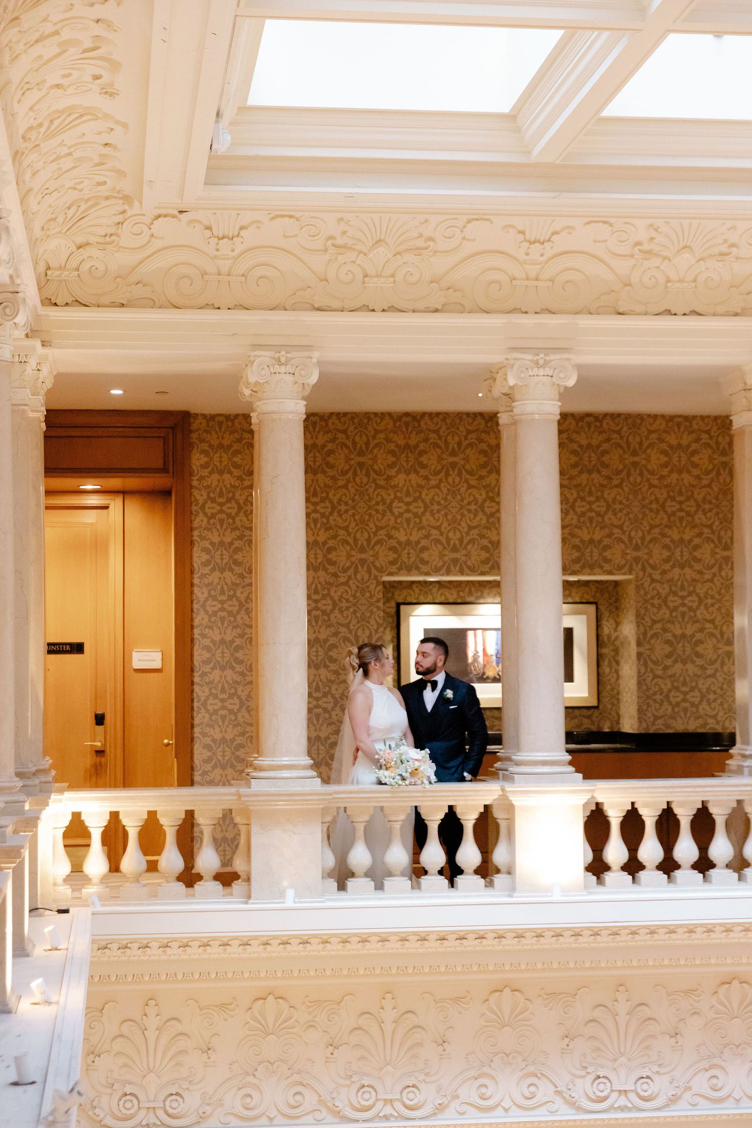 A grand interior balcony with white columns and ornate details showcases a wedding couple.