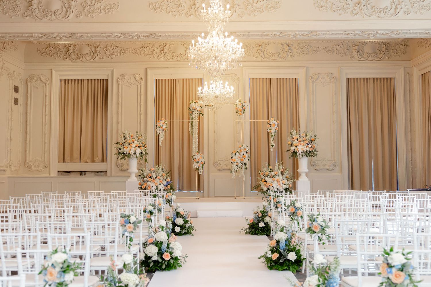 Elegant ballroom with crystal chandeliers, white chairs, and floral arrangements set up for a wedding ceremony.