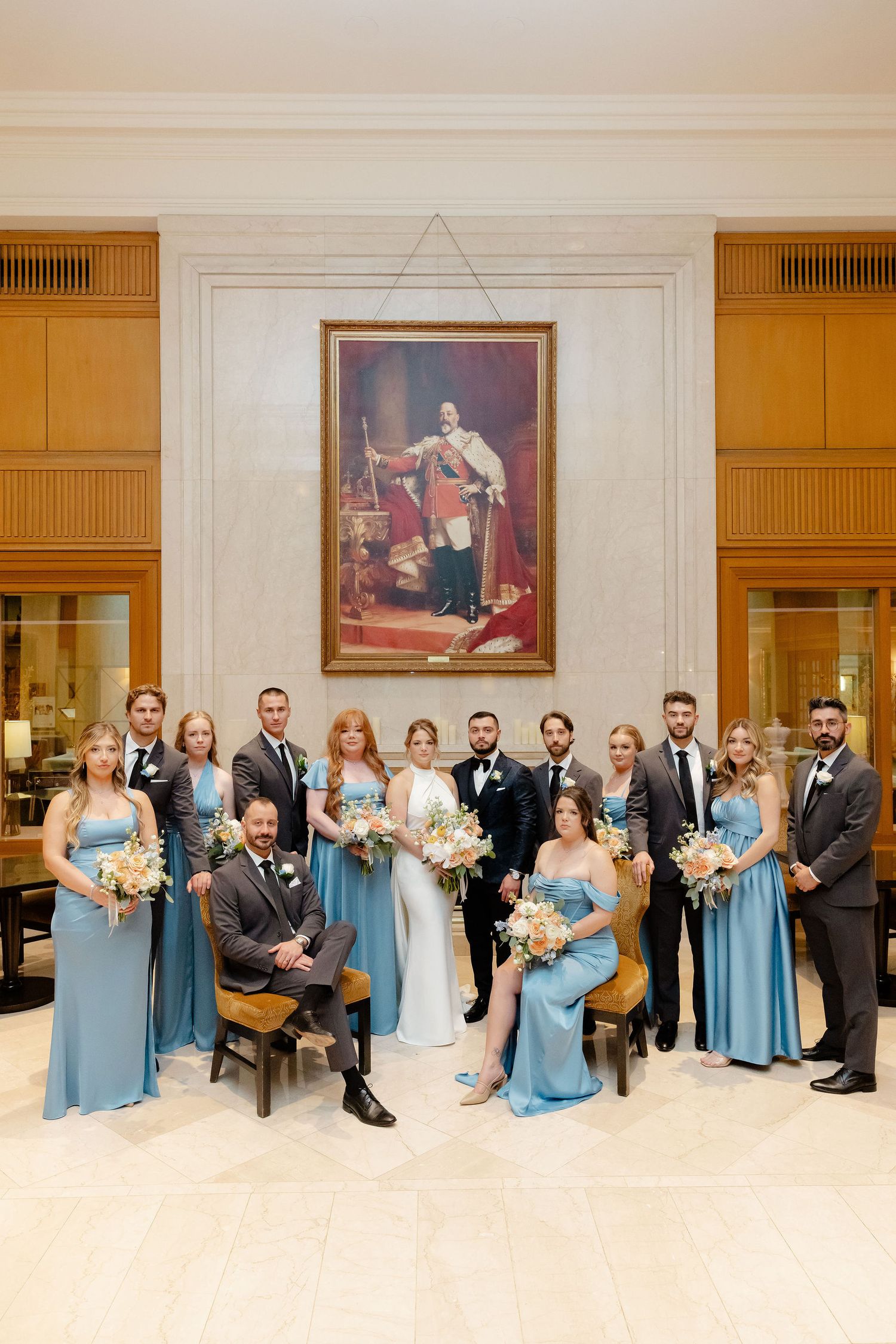Wedding party in blue dresses and dark suits poses together in an elegant venue with framed artwork.
