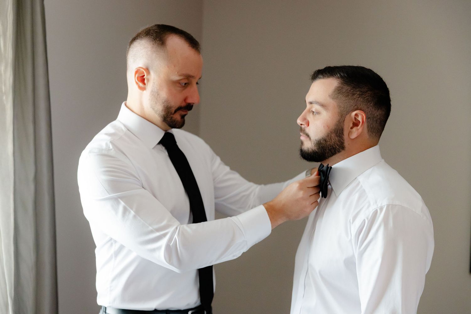Two people in white shirts and black ties help each other get dressed for a formal occasion.