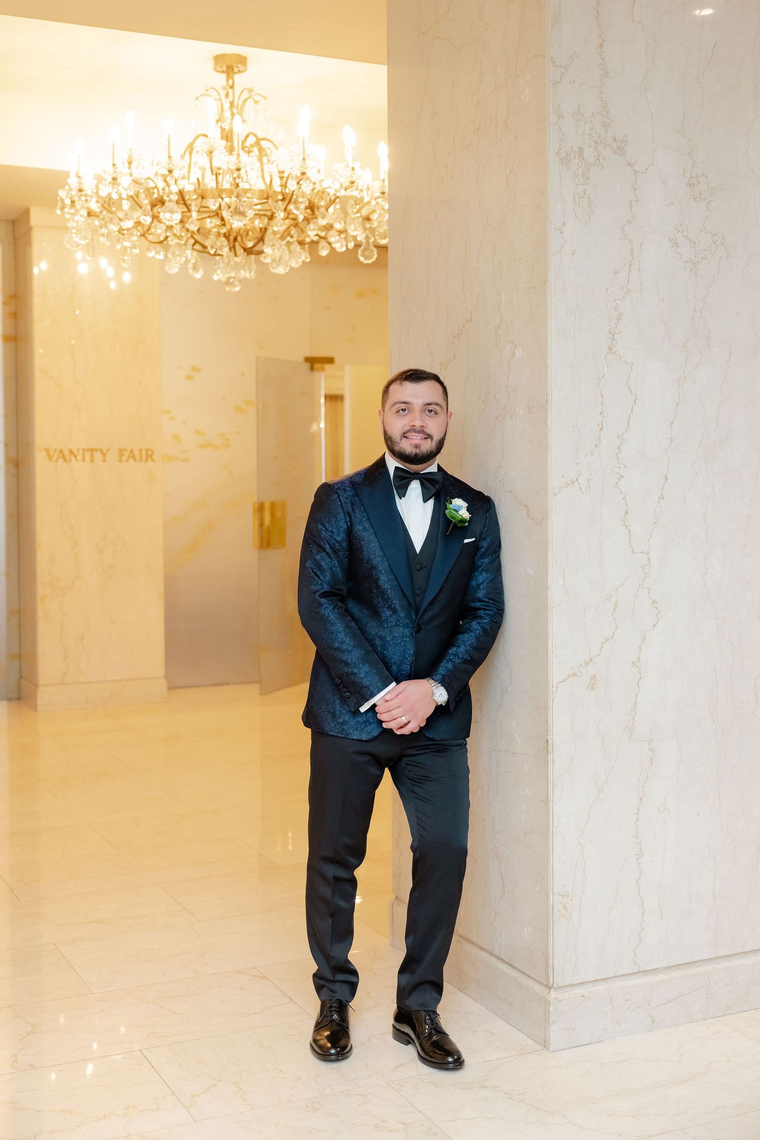 Groom in black tuxedo poses casually against marble columns in elegant venue.