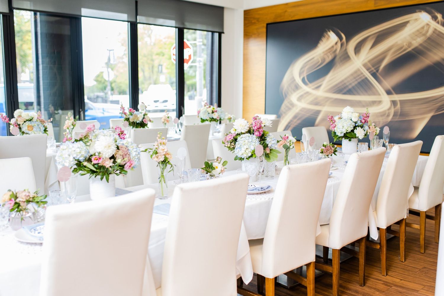 Elegant dining room setup with white chairs and floral centerpieces against a modern art wall installation.