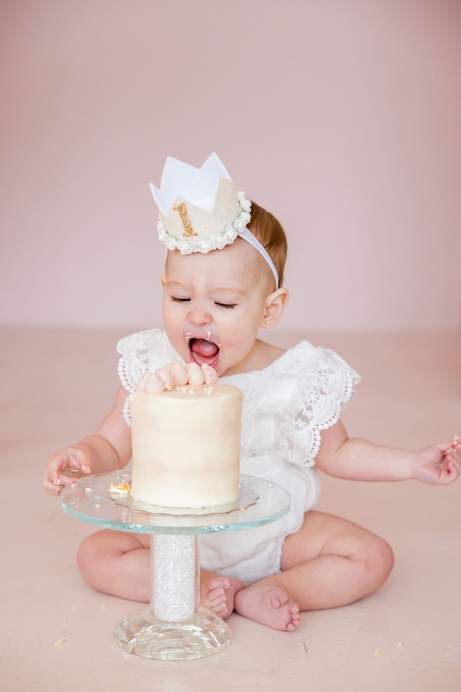 Baby in white dress and crown reaching for vanilla birthday cake on pink backdrop.