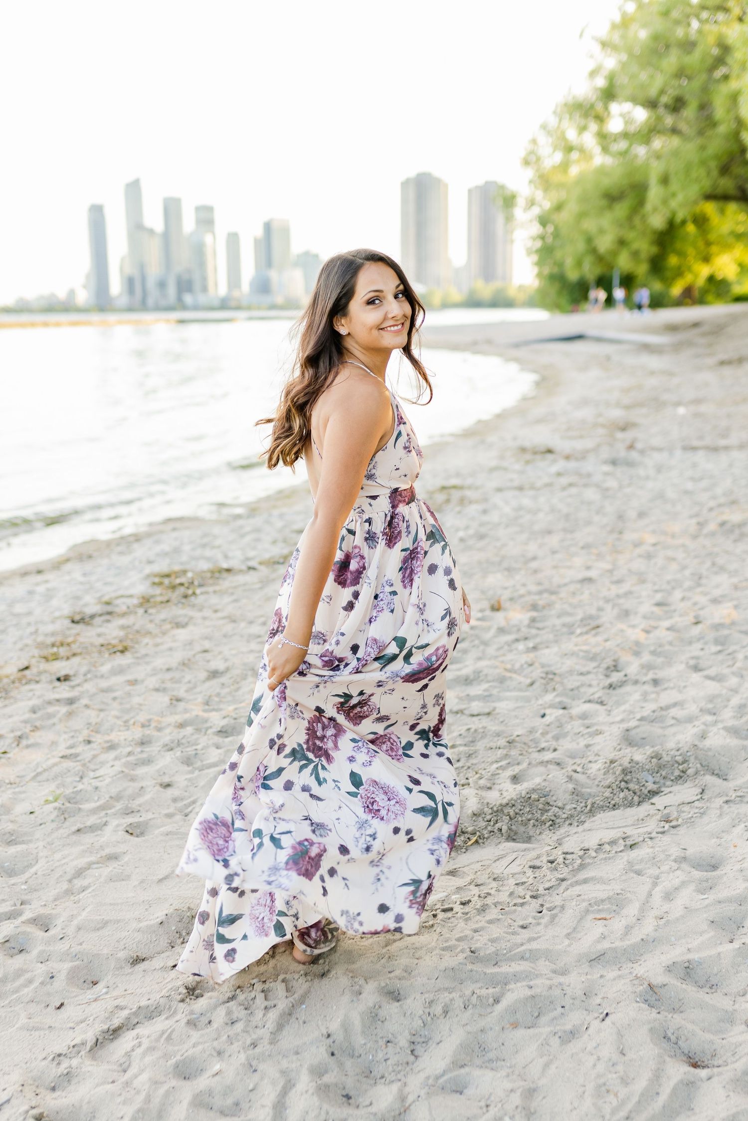 A woman in a flowing floral dress poses on a beach with city skyline in background.