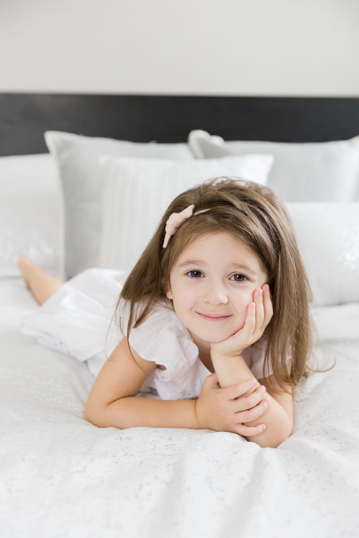 Child in white dress lying on bed with pillows and white bedding.