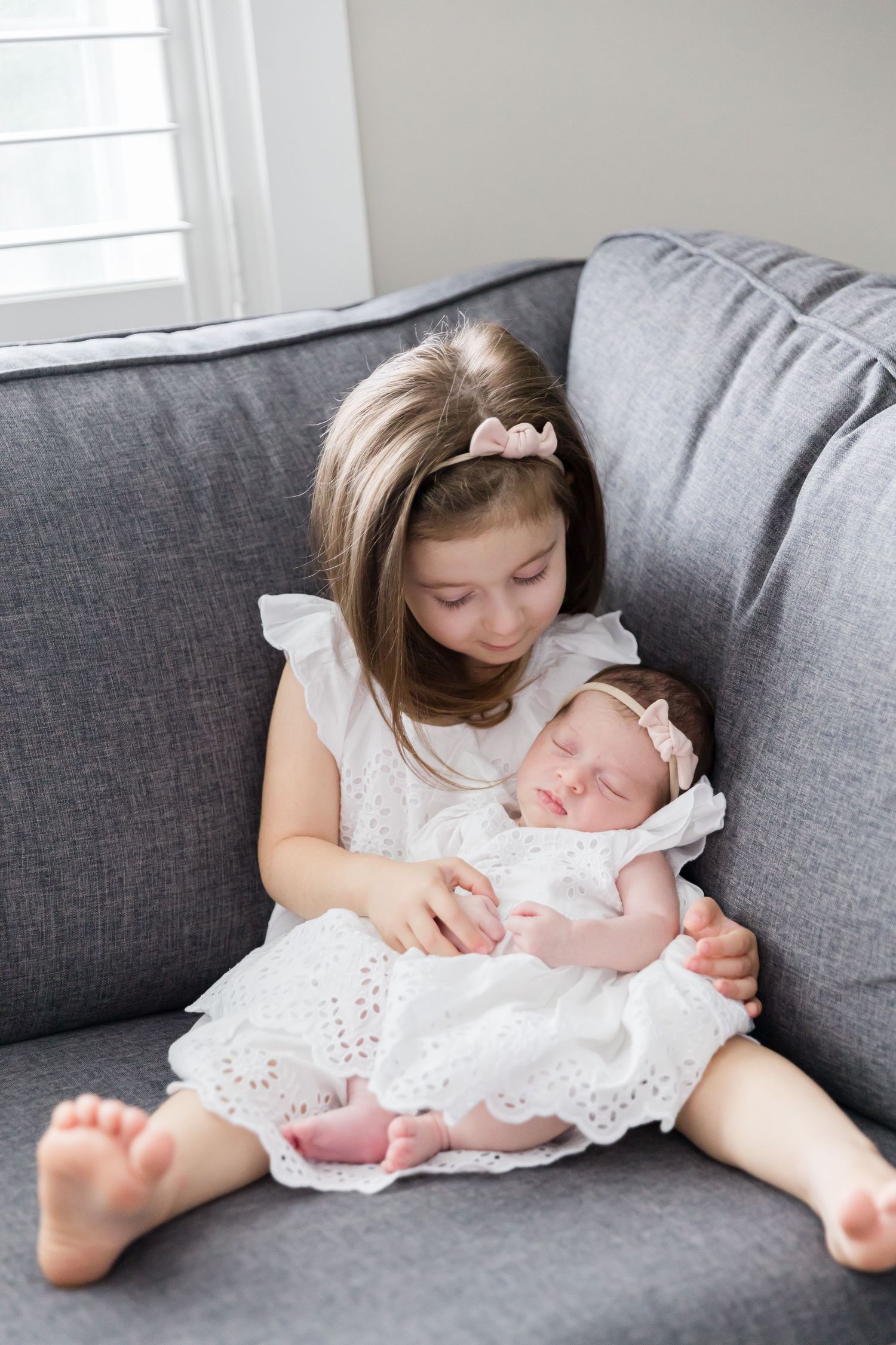 Child in white dress holds sleeping baby in matching white dress on grey couch.