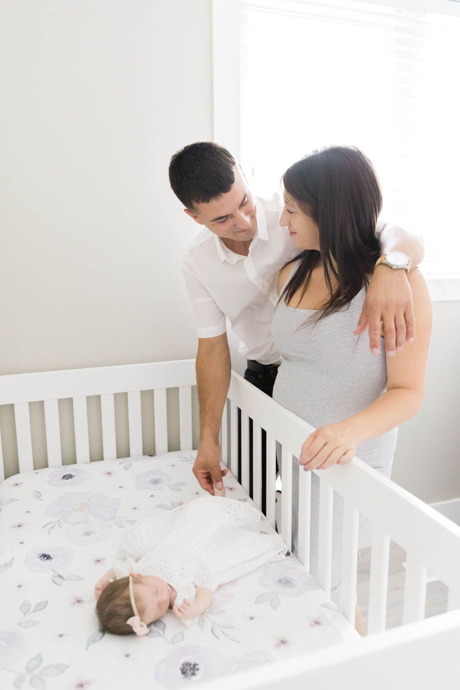 Couple standing by white crib watching their sleeping infant.