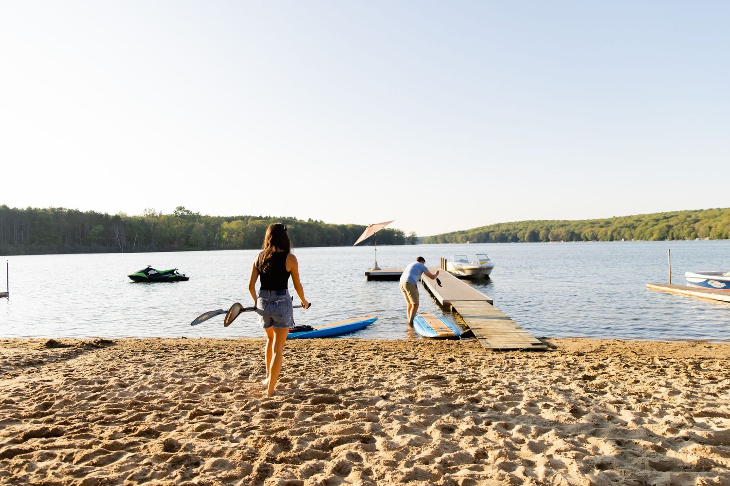 Two people walking along sandy beach near lake with docked boats in background.