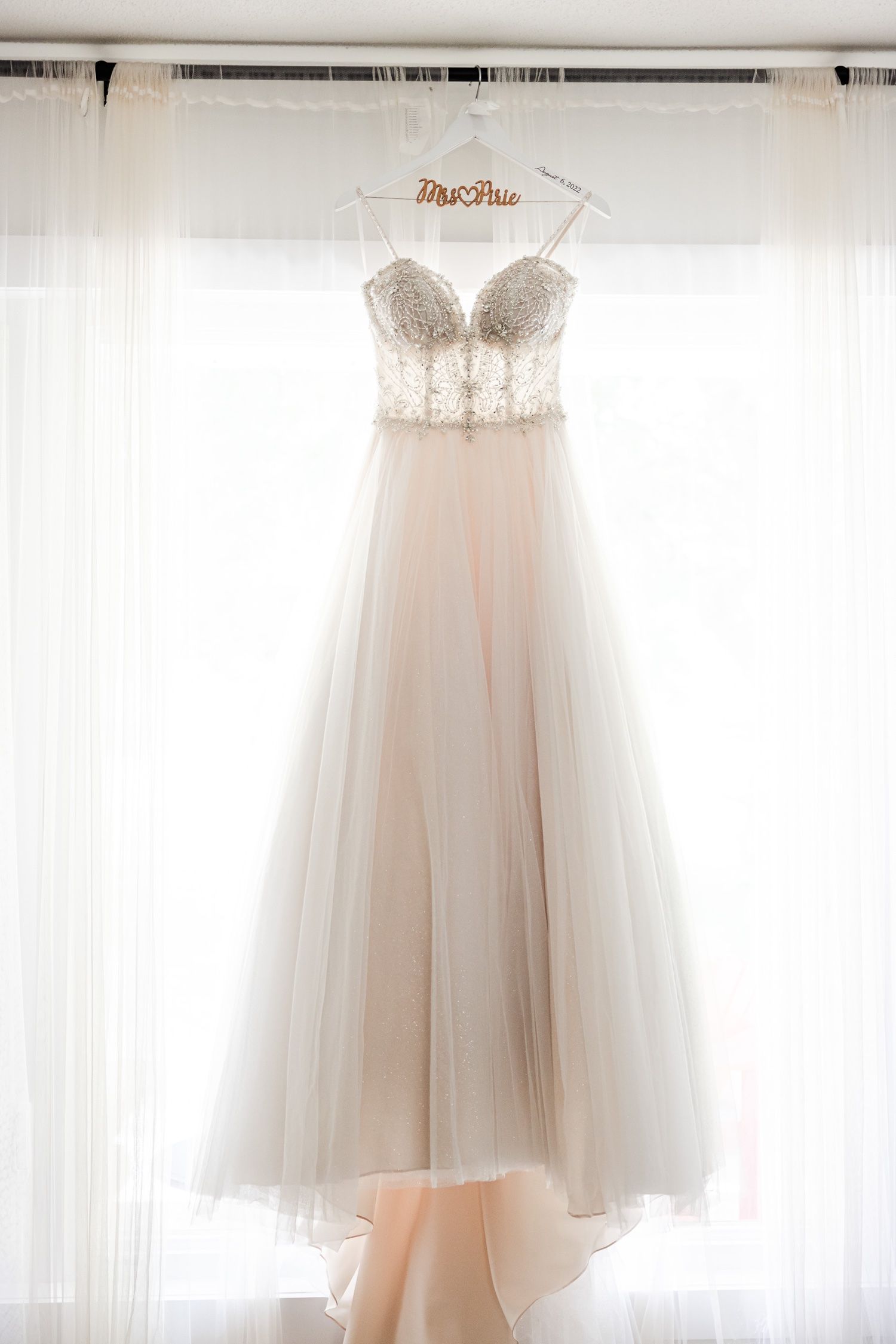 Beautiful white wedding dress hanging against sheer curtain backdrop.