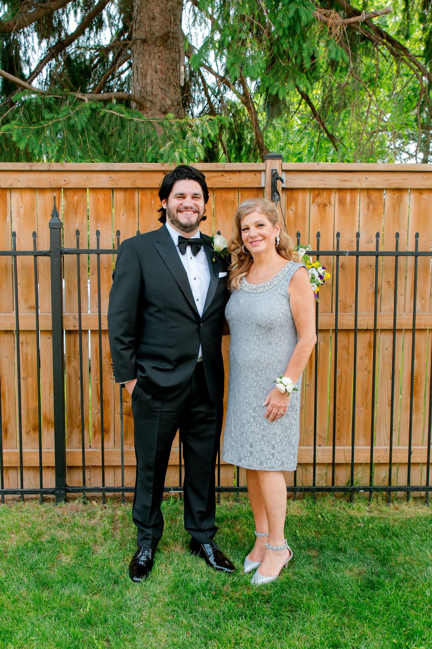 Formal couple poses in front of wooden fence at outdoor event, wearing black tuxedo and silver cocktail dress.