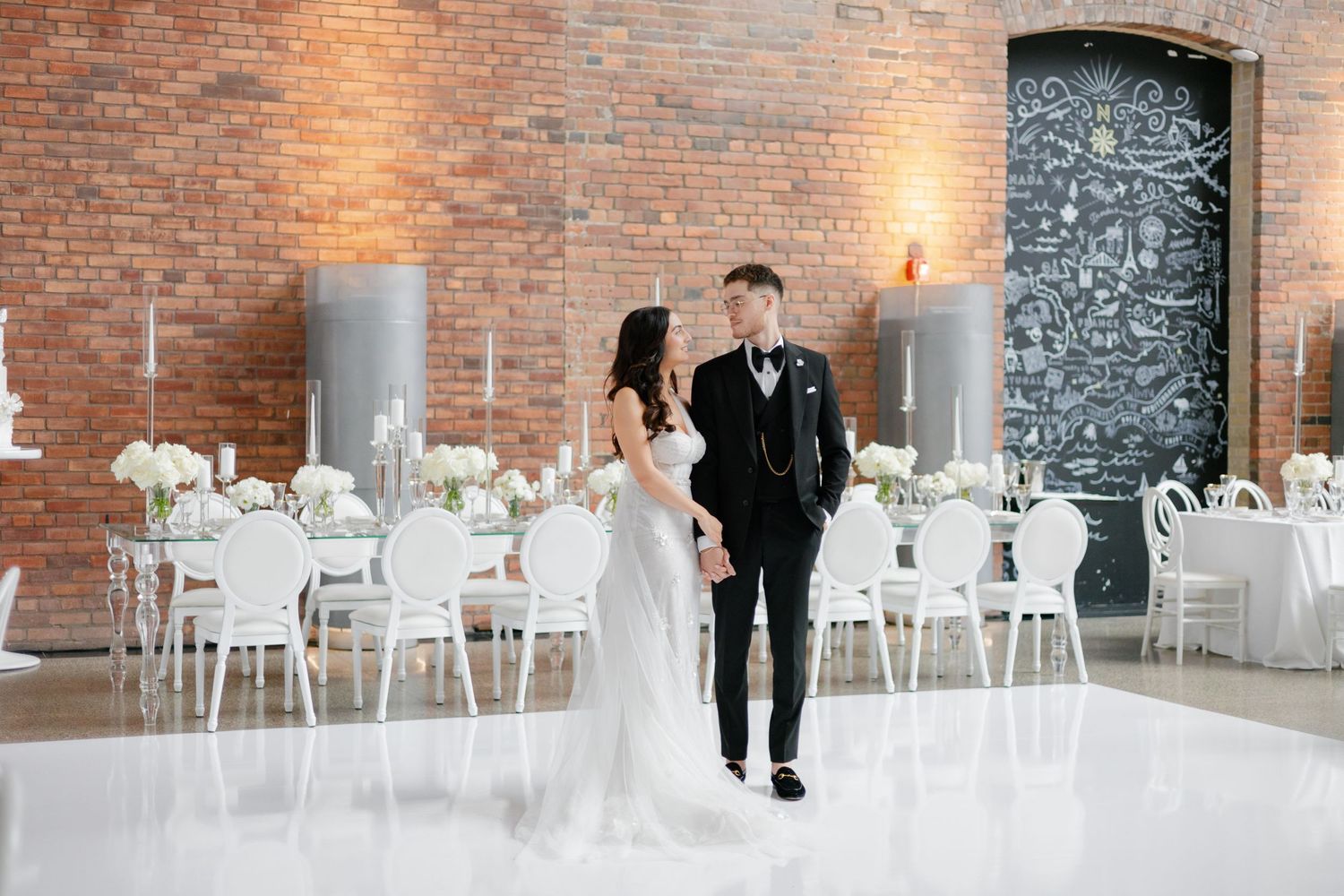 Wedding couple walking in a modern reception space with exposed brick walls and white decor.