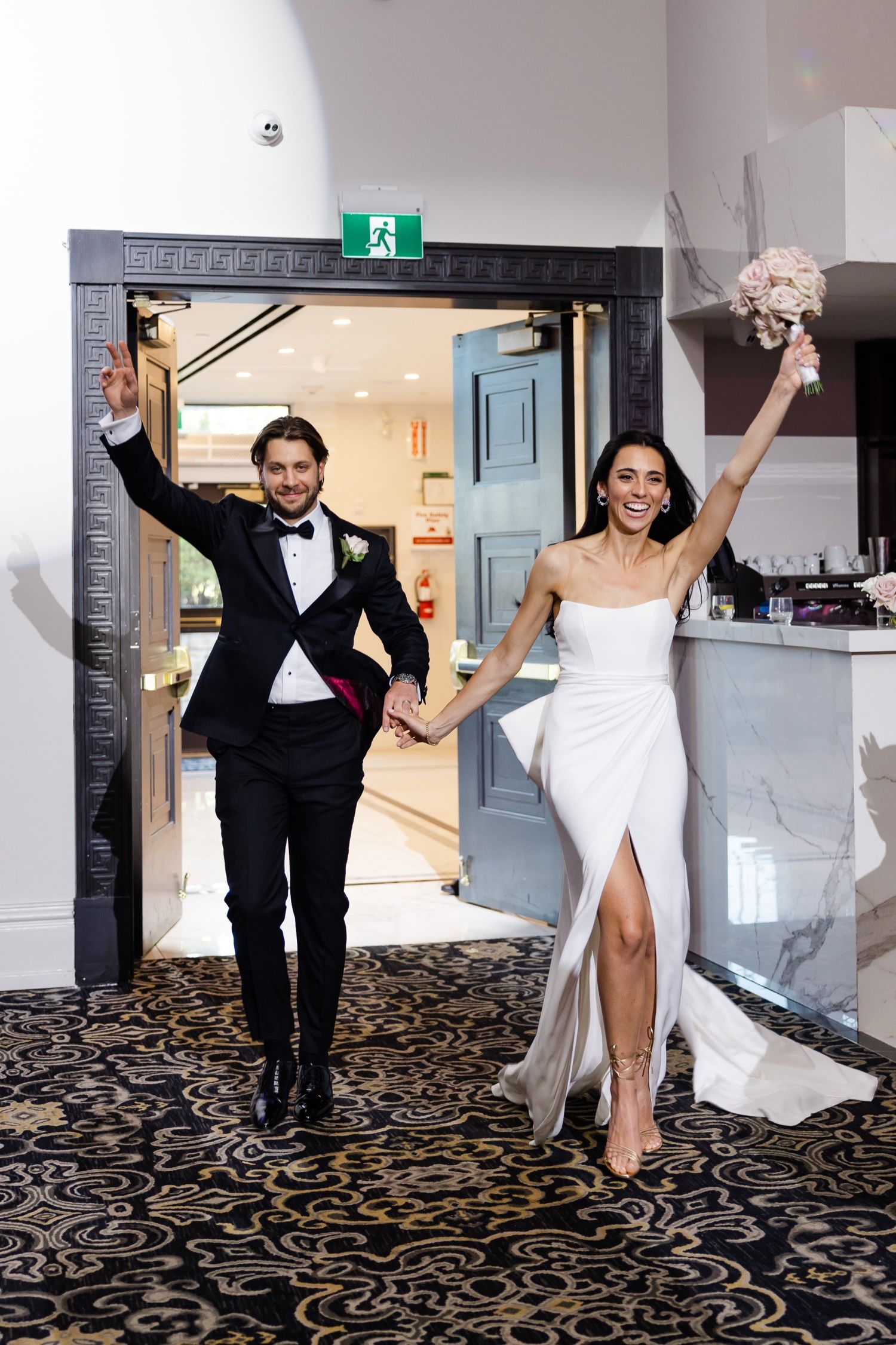 Newlyweds celebrating with arms raised as they enter through decorated doorway.