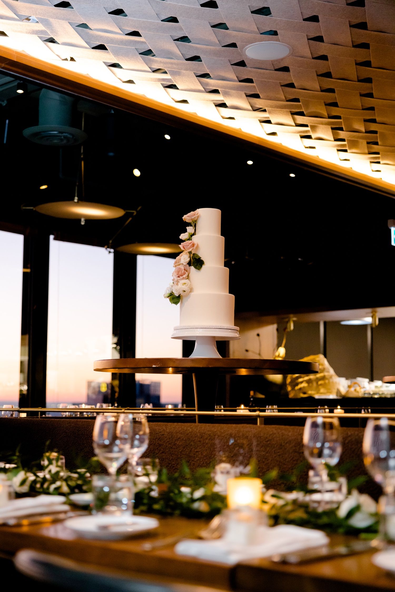 White tiered wedding cake displayed against sunset view through large windows at reception venue.