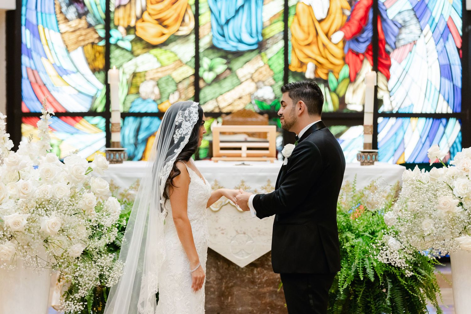 A couple exchanging vows in front of a colorful stained glass window with white floral decorations.