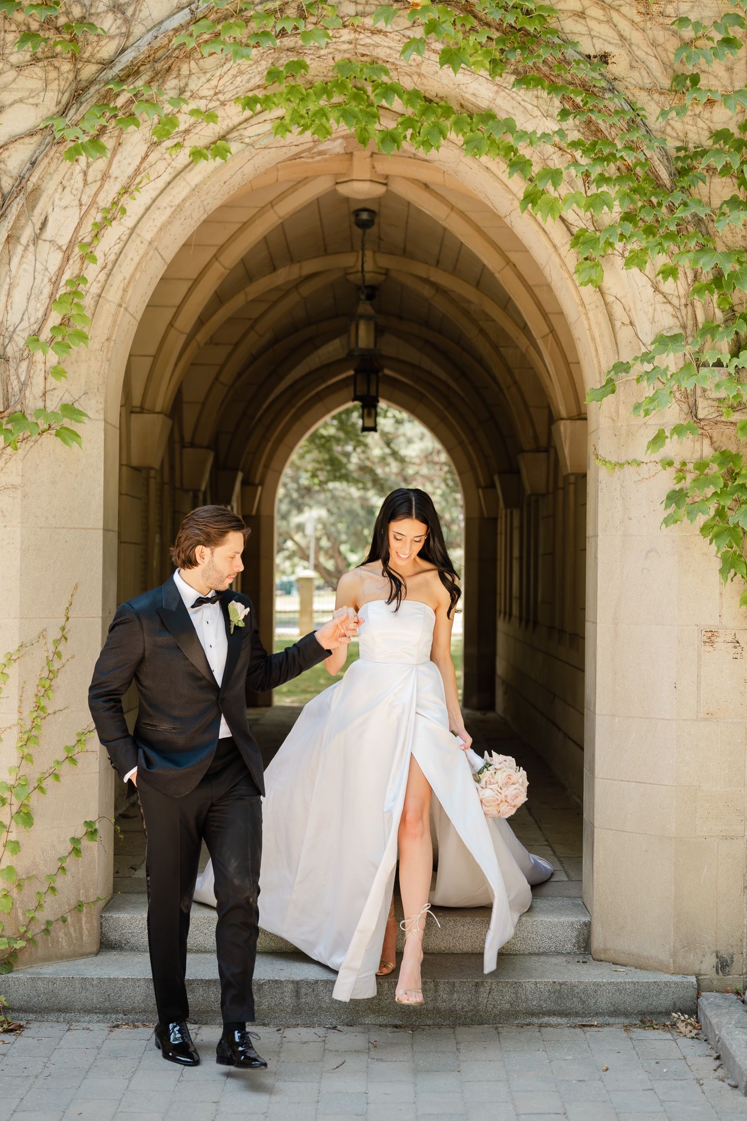 A romantic wedding couple stands in a sunlit stone archway with climbing vines.