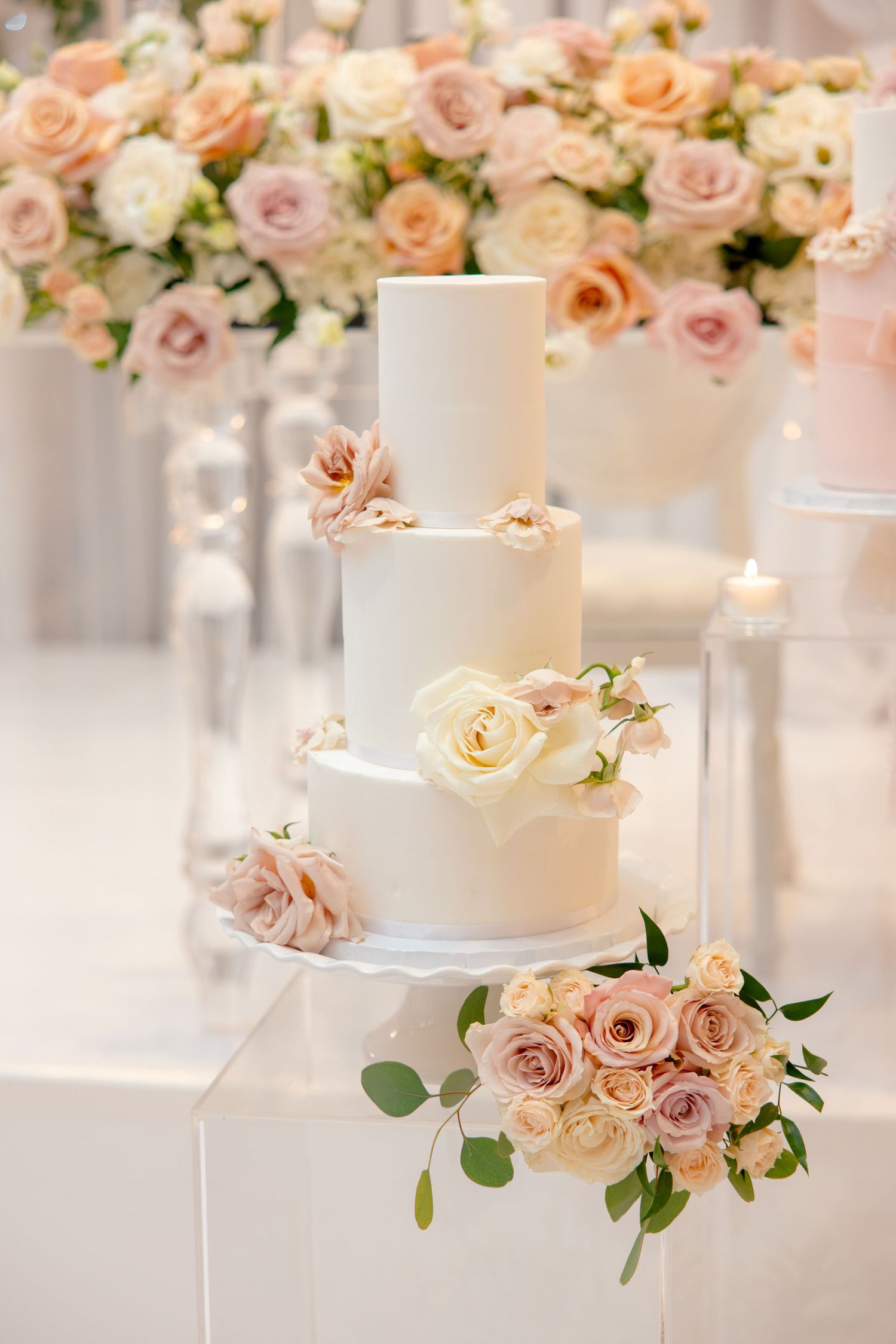 Three-tiered white wedding cake decorated with blush and peach roses against a floral backdrop.