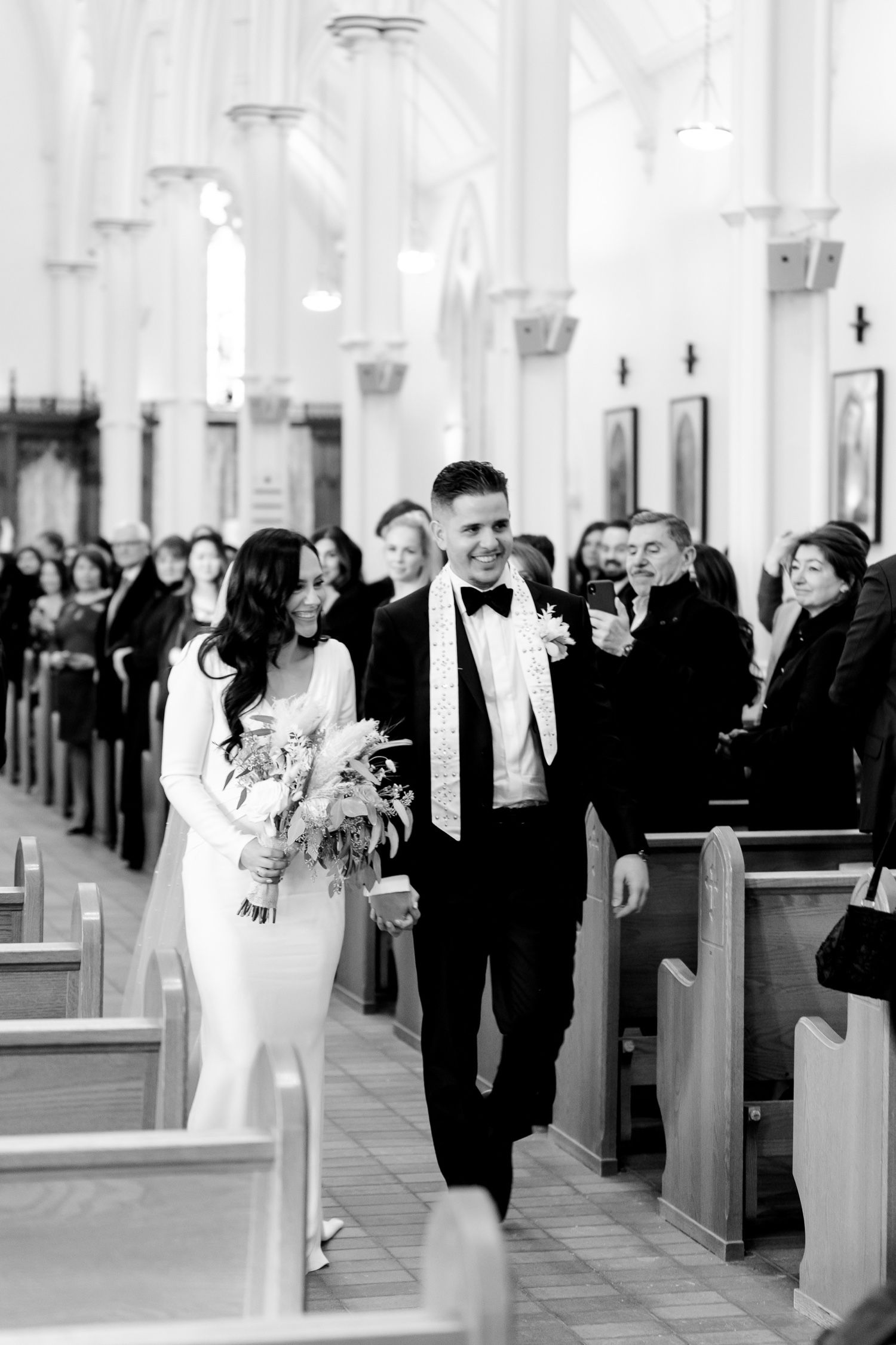 A black and white photo of newlyweds walking down the church aisle with smiling guests.