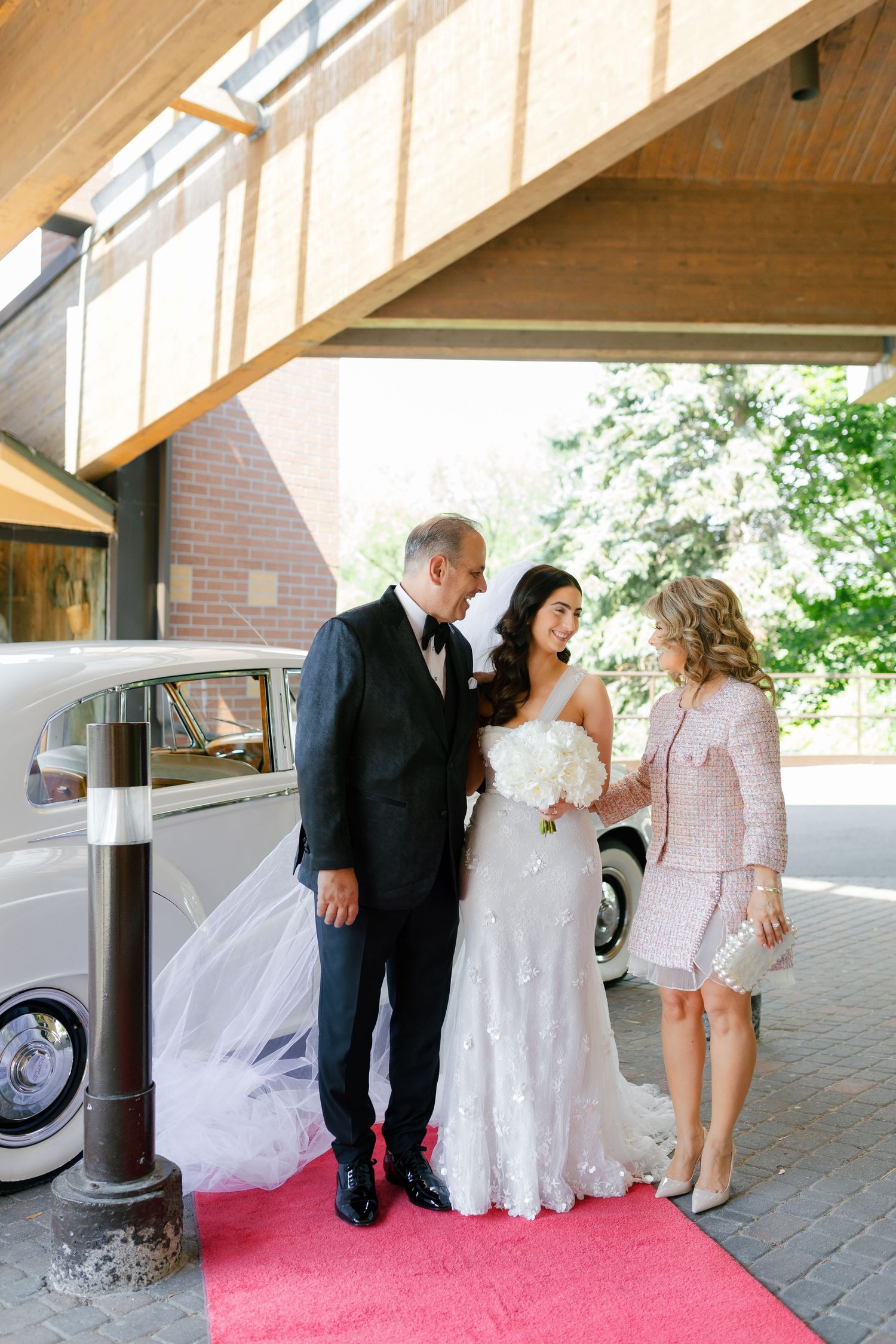 Wedding party members pose together on a red carpet outside with a classic white car in the background.