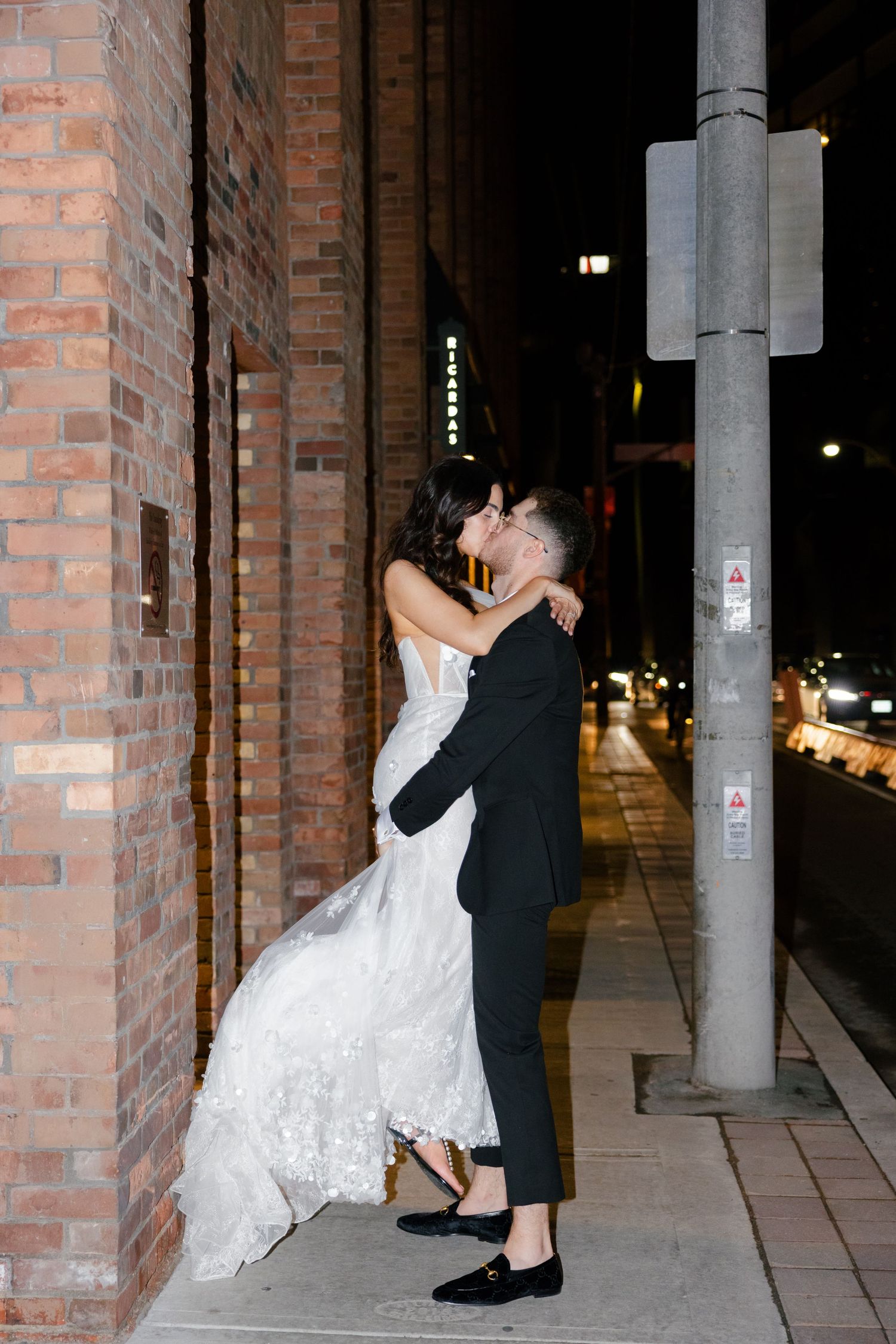 Romantic nighttime moment between newlyweds against urban brick wall setting.