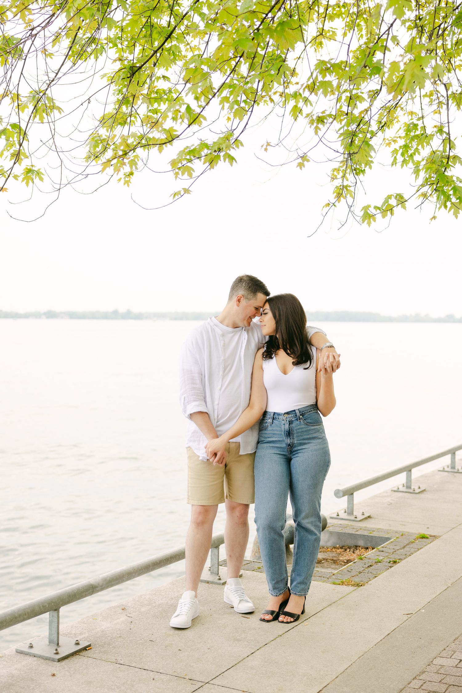 A couple embracing on a waterfront walkway under green spring foliage.
