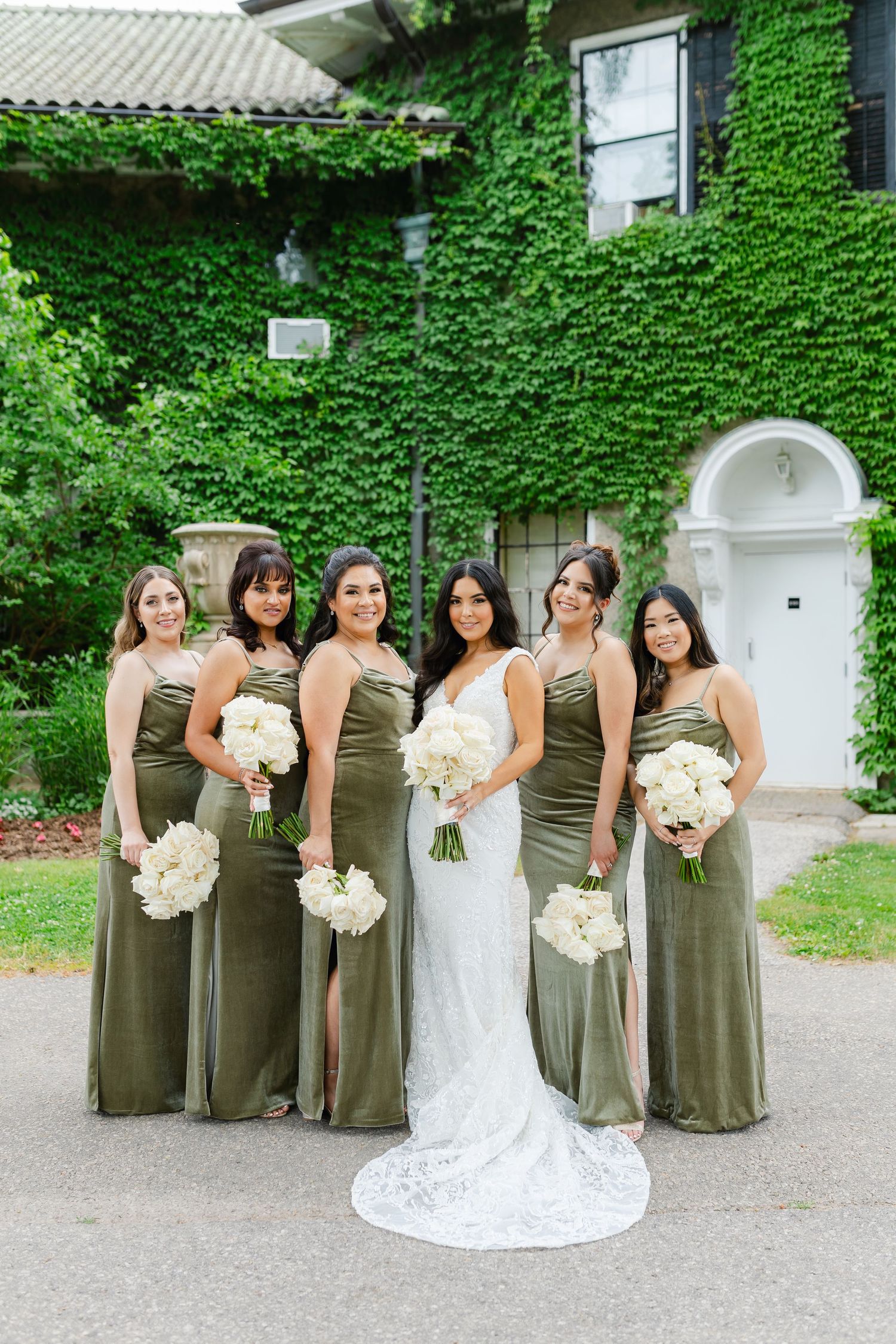 Wedding party in olive green dresses poses with white floral bouquets against historic building.