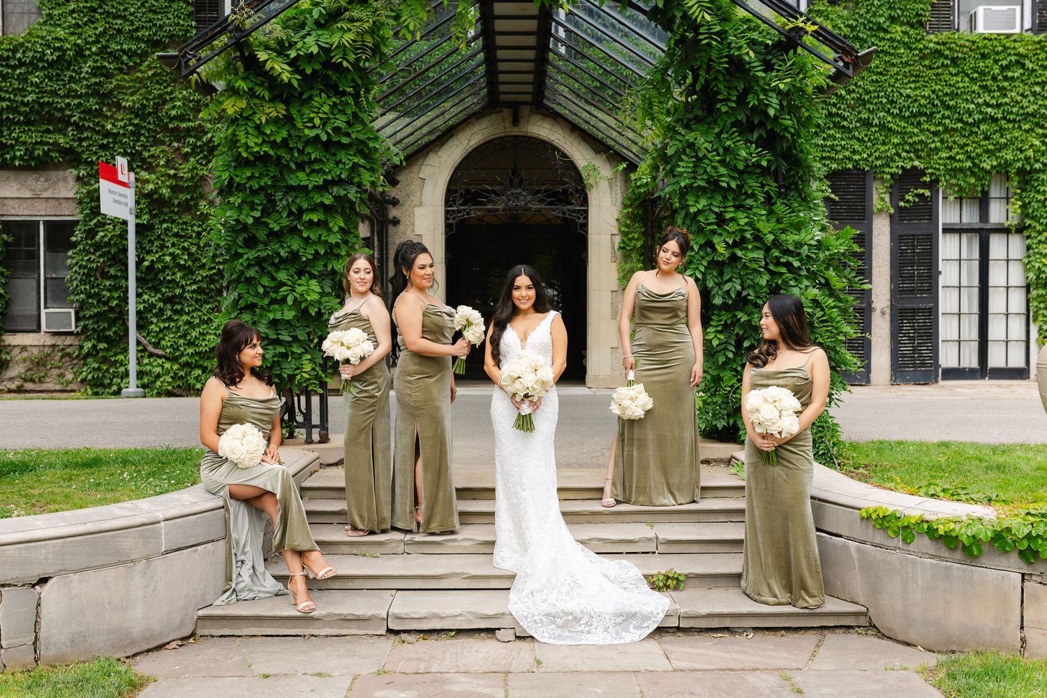 Wedding party in sage green dresses and white gown pose on stone steps of ivy-covered archway.