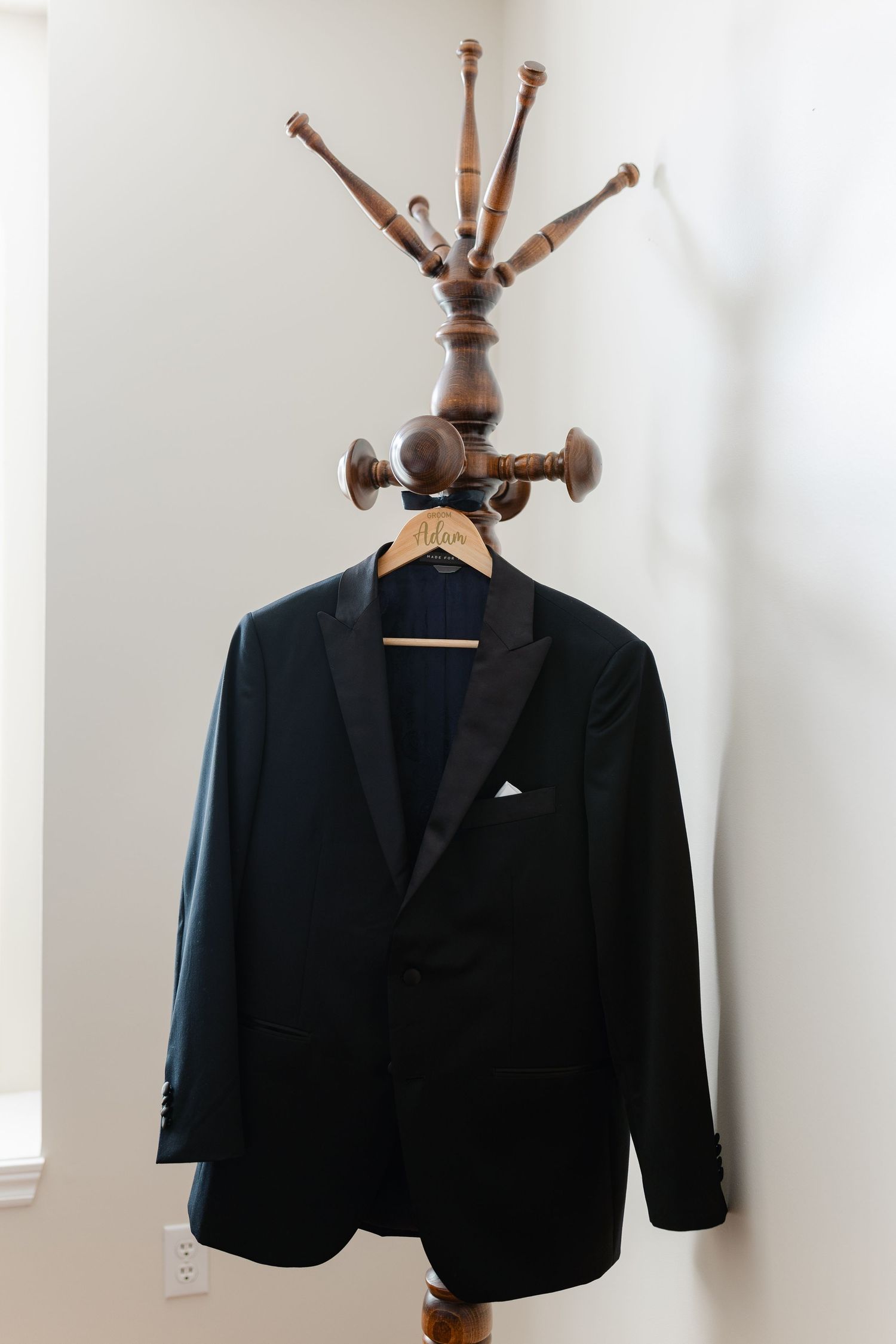 A black tuxedo jacket hung on an antique wooden coat stand against a white wall.