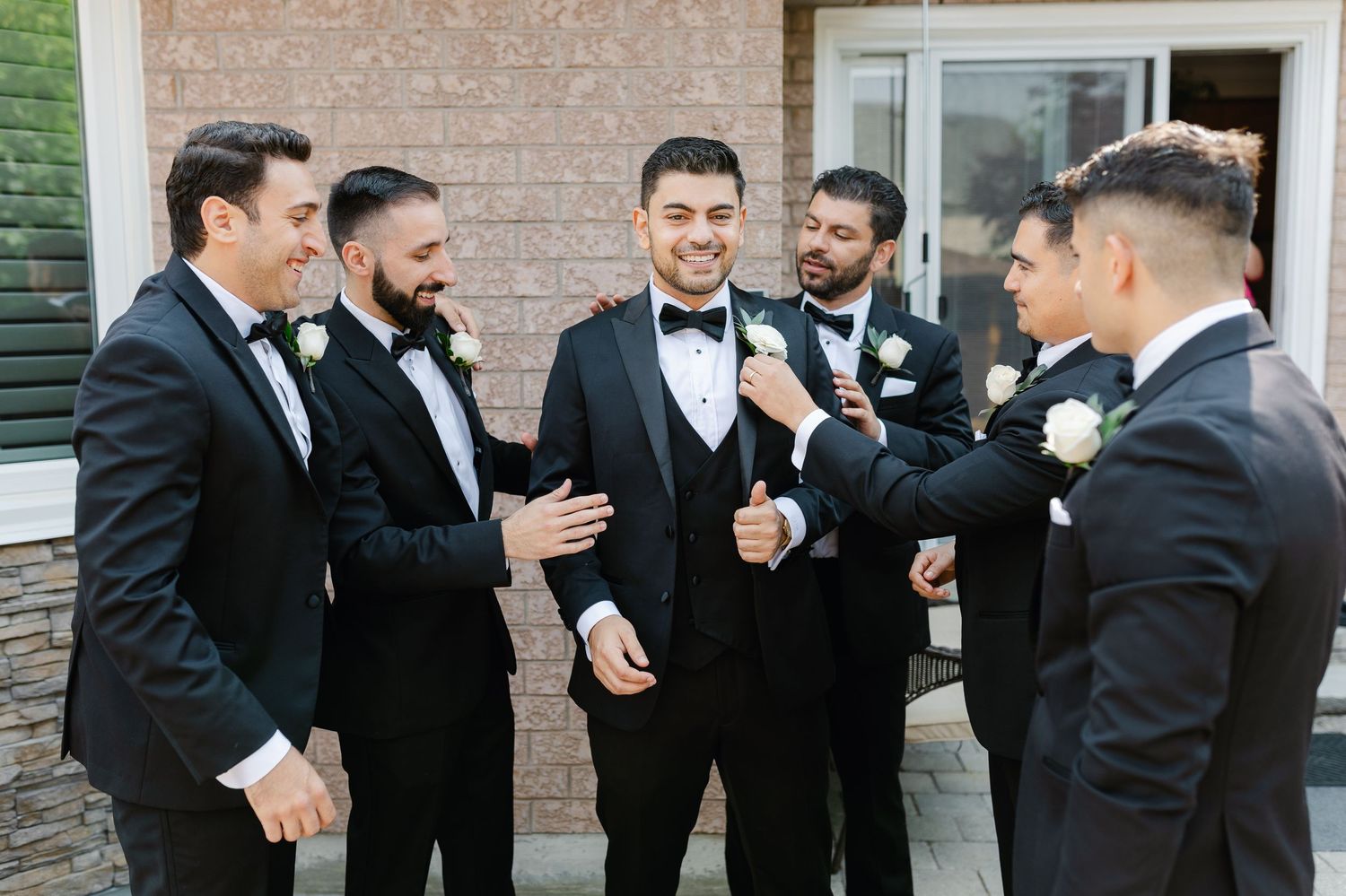 Group of groomsmen in black tuxedos share laughs during outdoor wedding photo session.