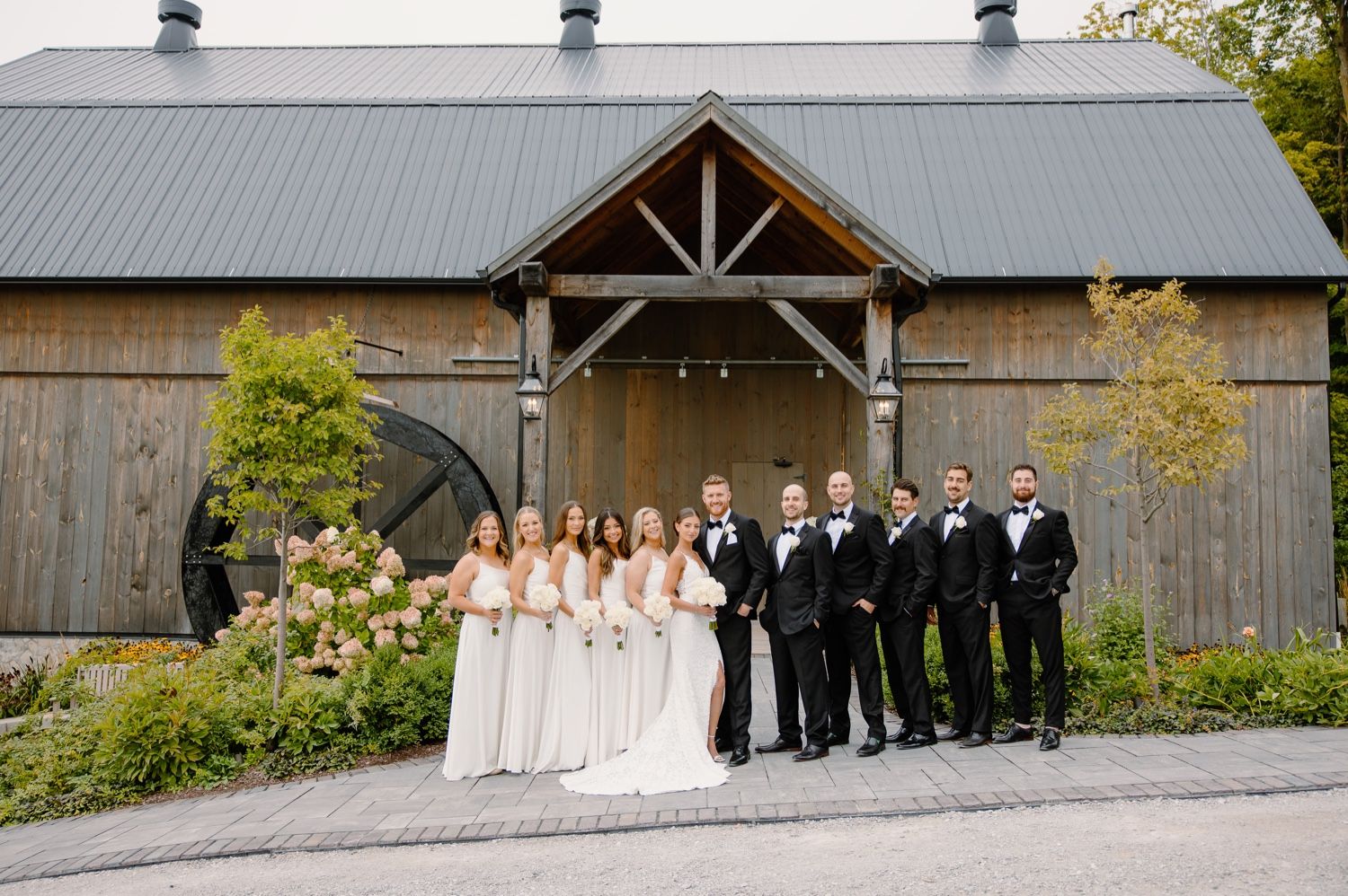 Full wedding party in formal attire posed together outside a rustic barn venue.