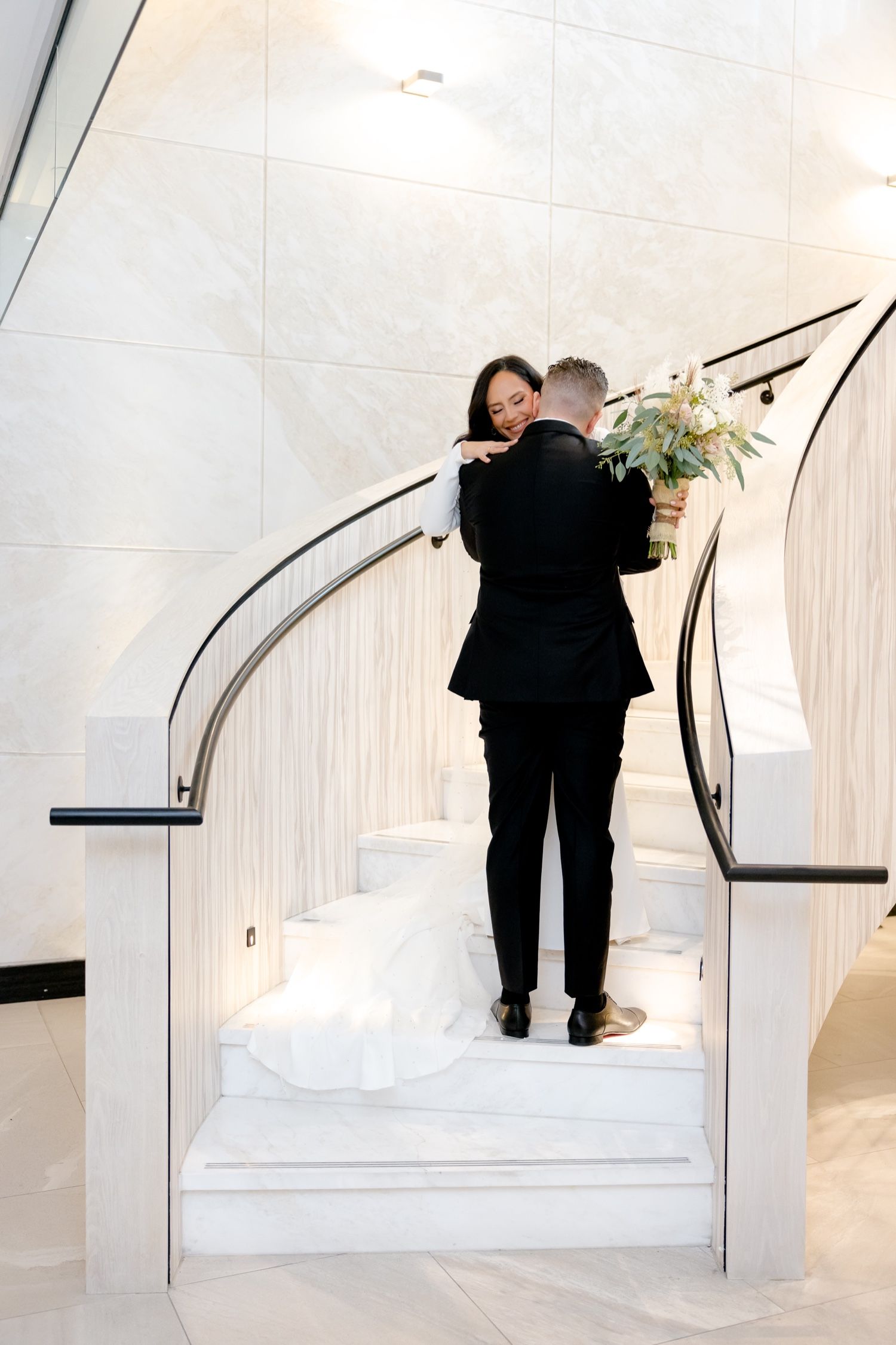 A tender embrace between newlyweds on white marble stairs with elegant curved railings.