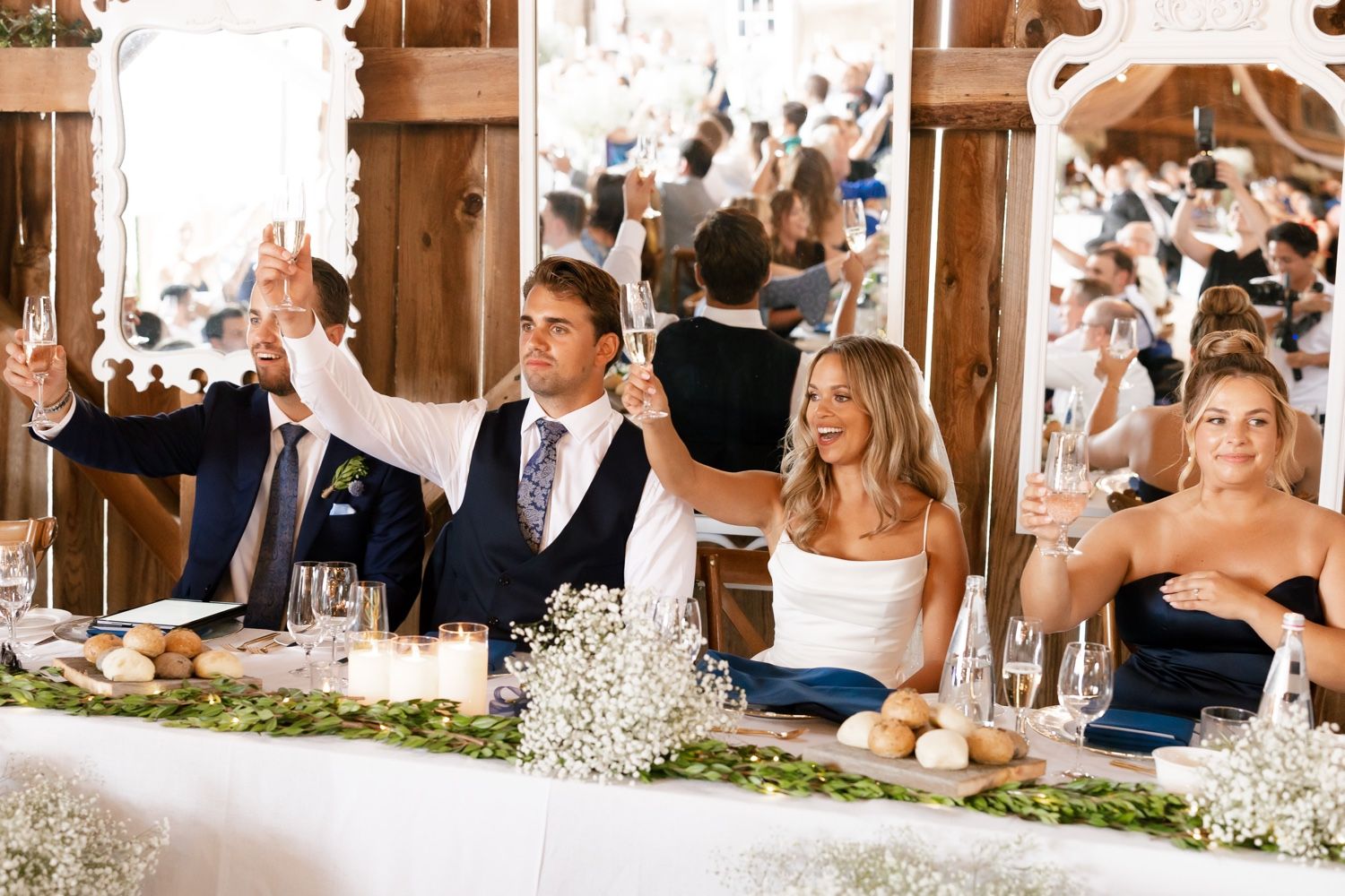 Wedding party members seated at head table raise their glasses in celebration during reception toasts.