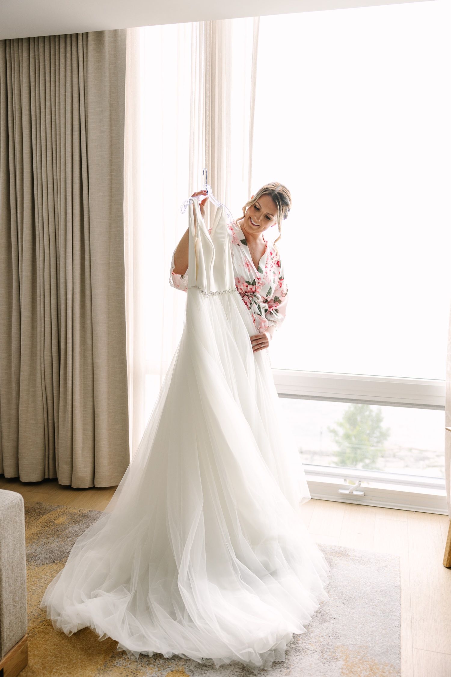 Wedding dress displayed in sunlit room with flowing tulle skirt and sheer curtains.