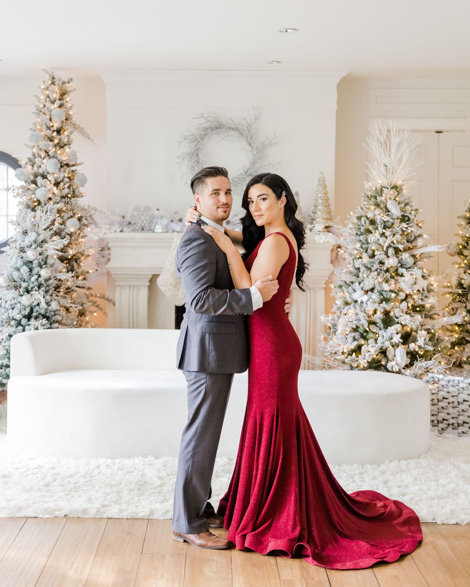 Elegant couple embraces in front of decorated Christmas trees in a bright white room.