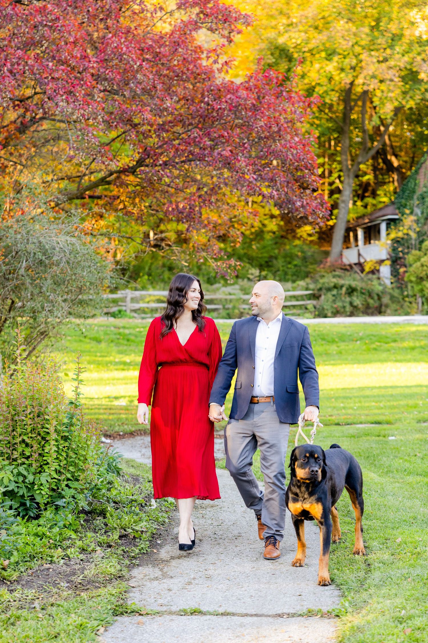 A couple walking their Rottweiler dog along a garden path surrounded by vibrant autumn foliage.