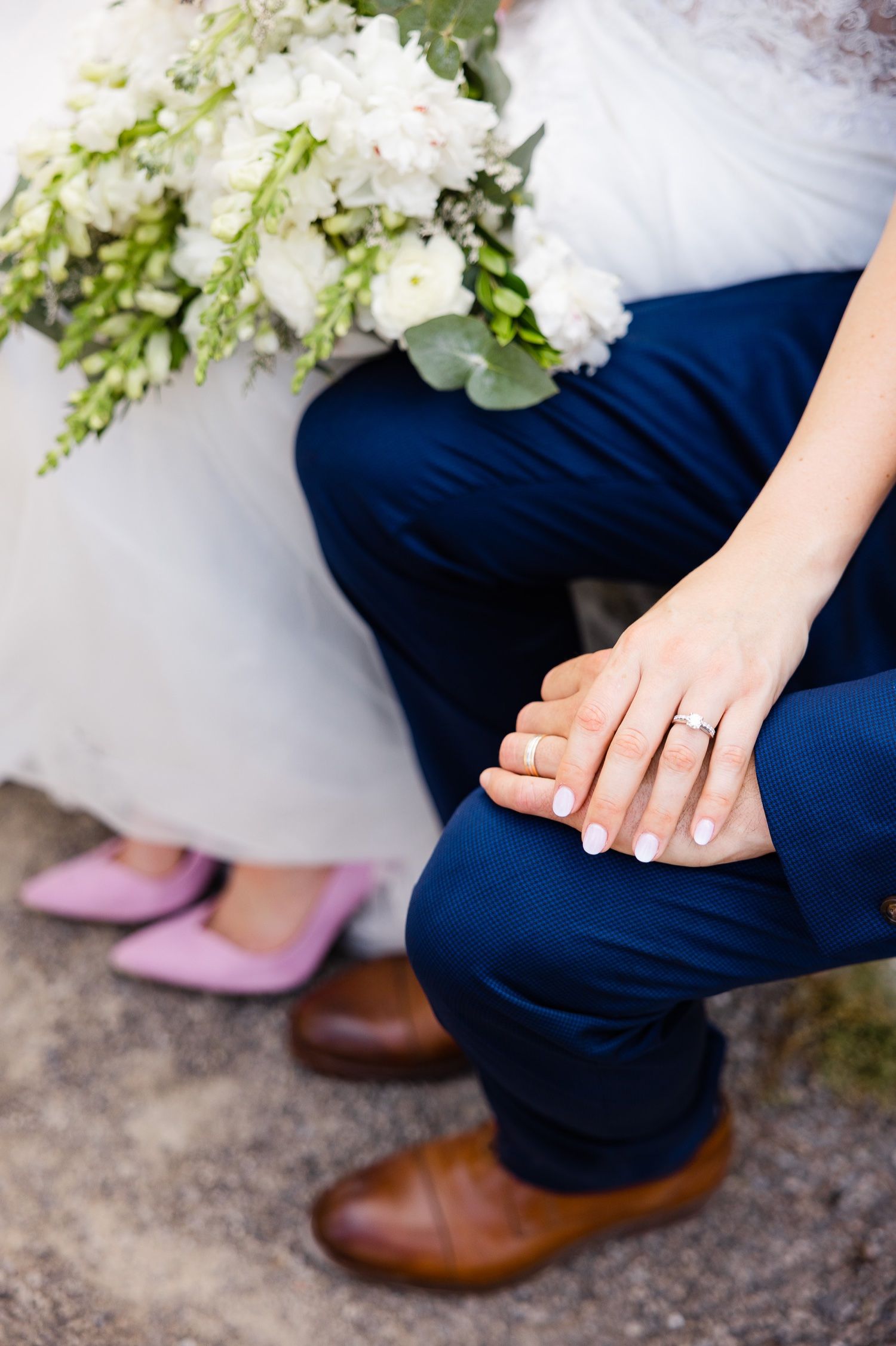 Close-up of wedding couple's hands showing ring with pink shoes, brown shoes and white bouquet visible.